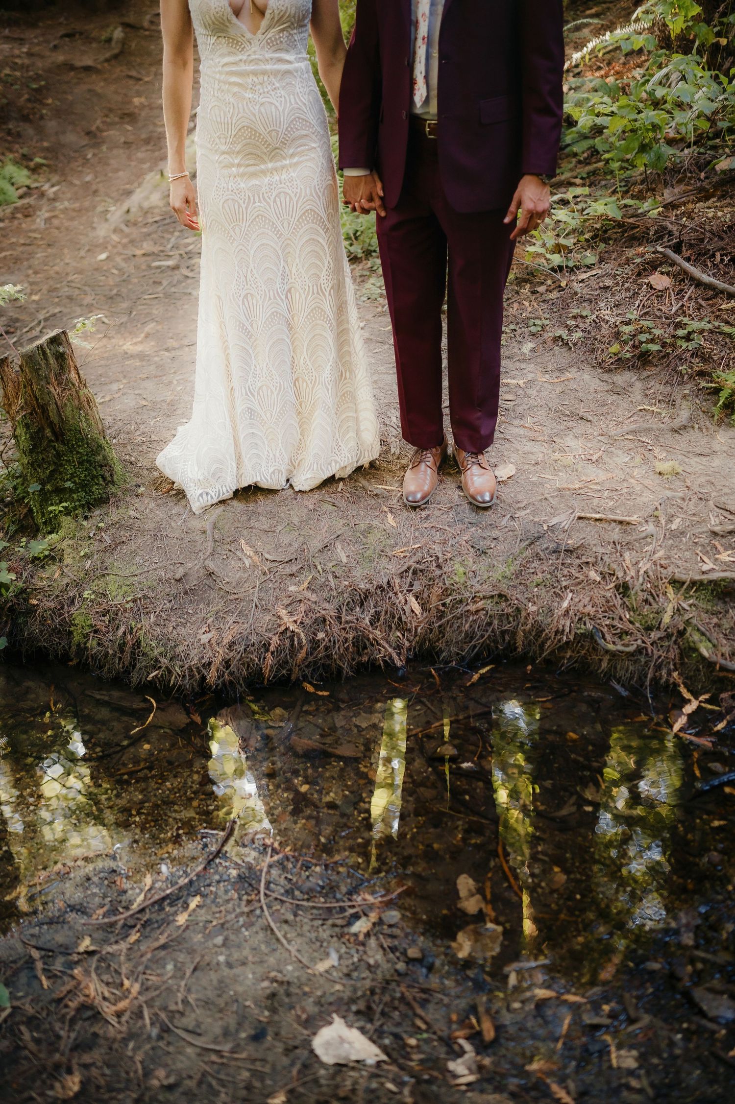 A romantic wedding couple standing near a reflective pond in a woodland setting.