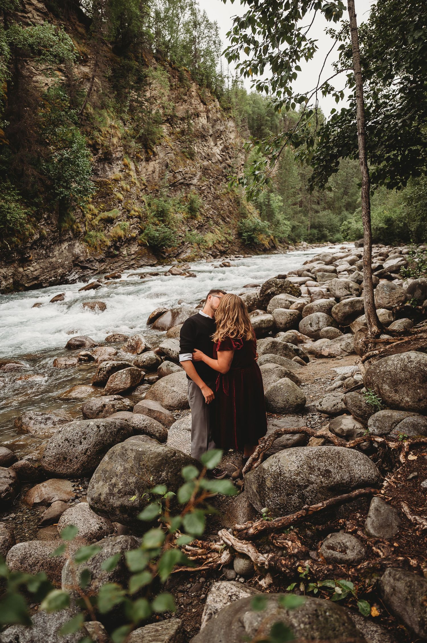 A romantic moment unfolds by a rushing river amidst rocky terrain and natural scenery.
