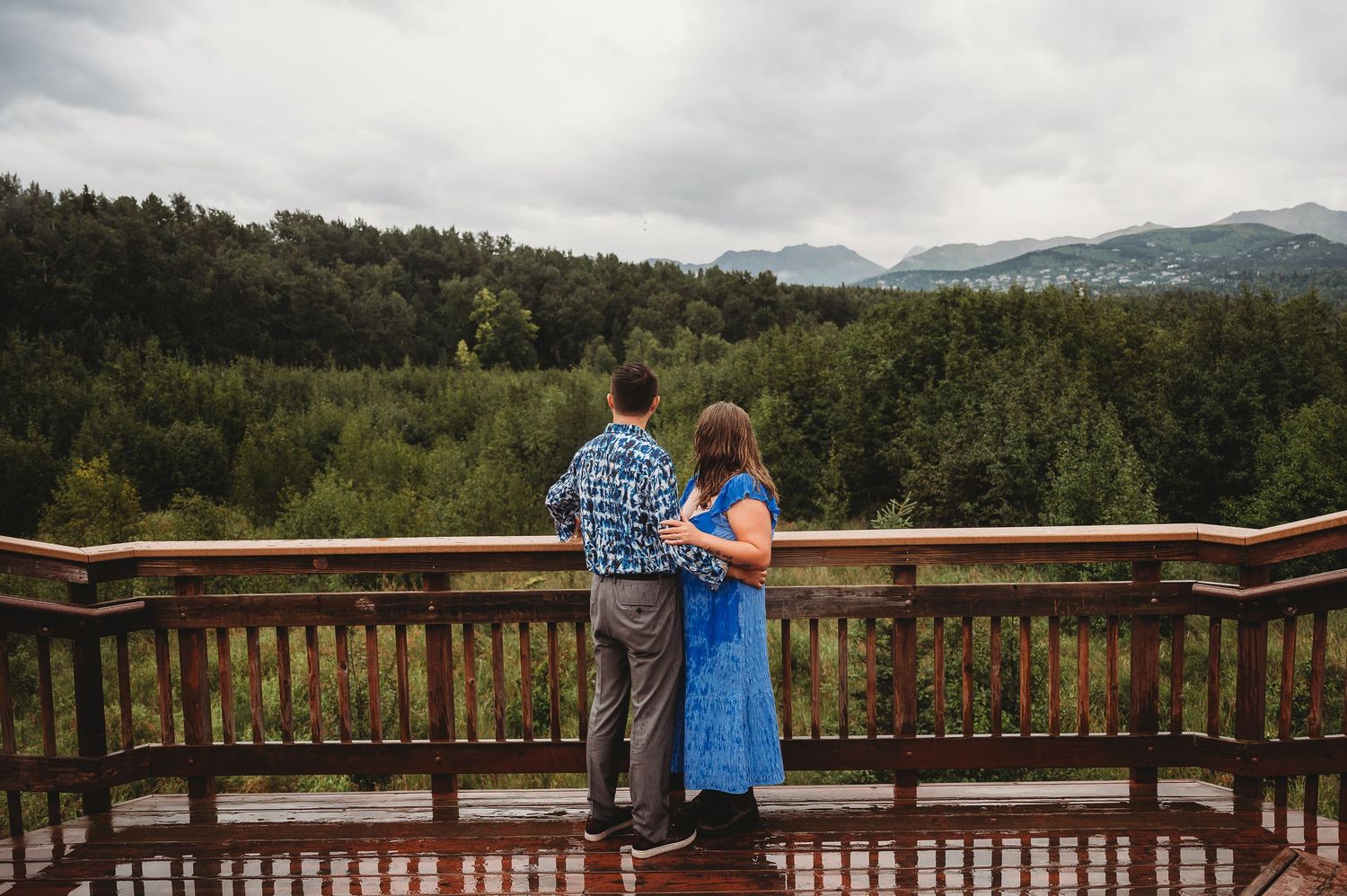 Two people stand on a wooden viewing deck overlooking a scenic mountain forest landscape under cloudy skies.