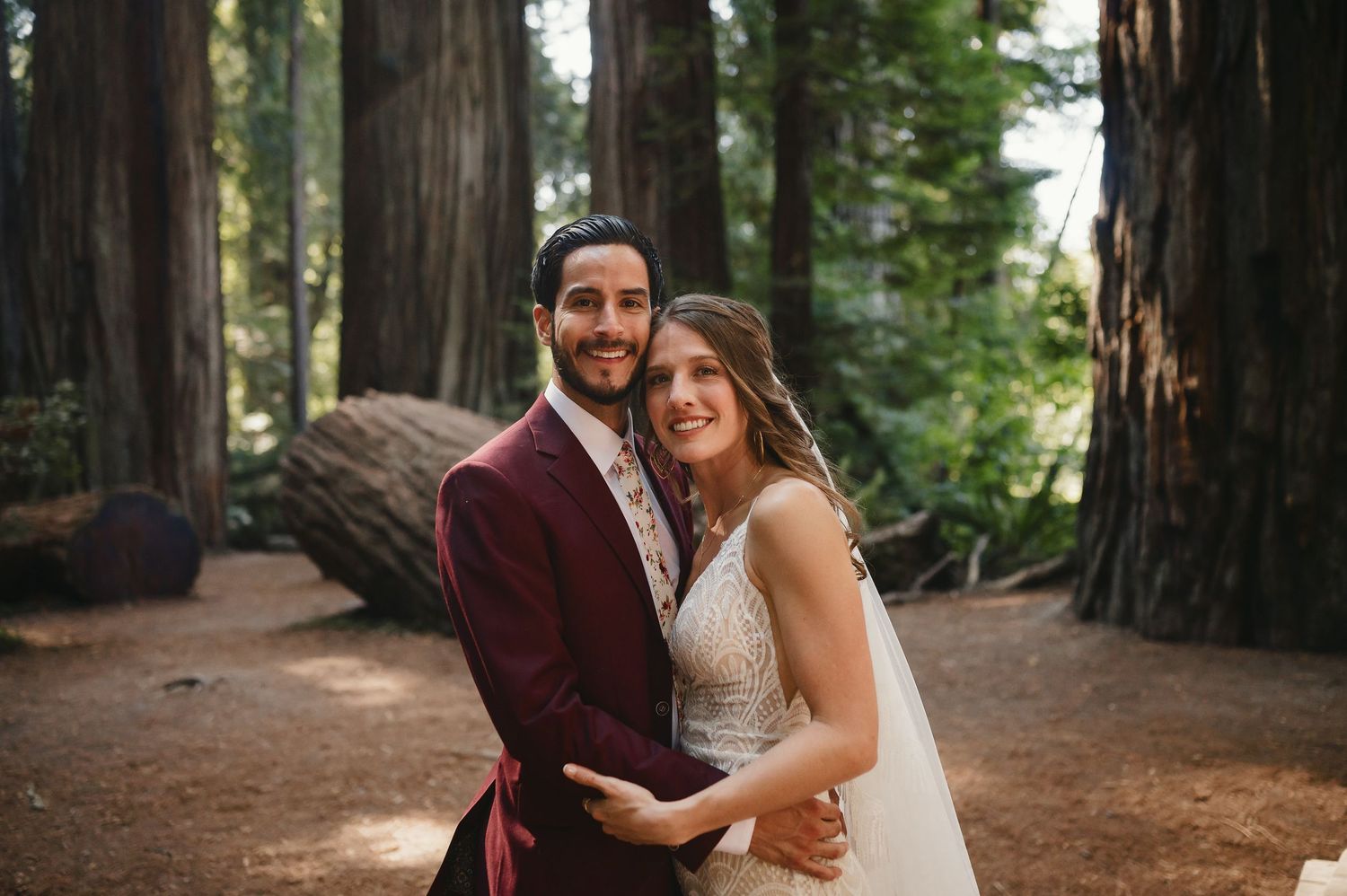Happy couple poses together in a redwood forest surrounded by towering trees along a dirt path.