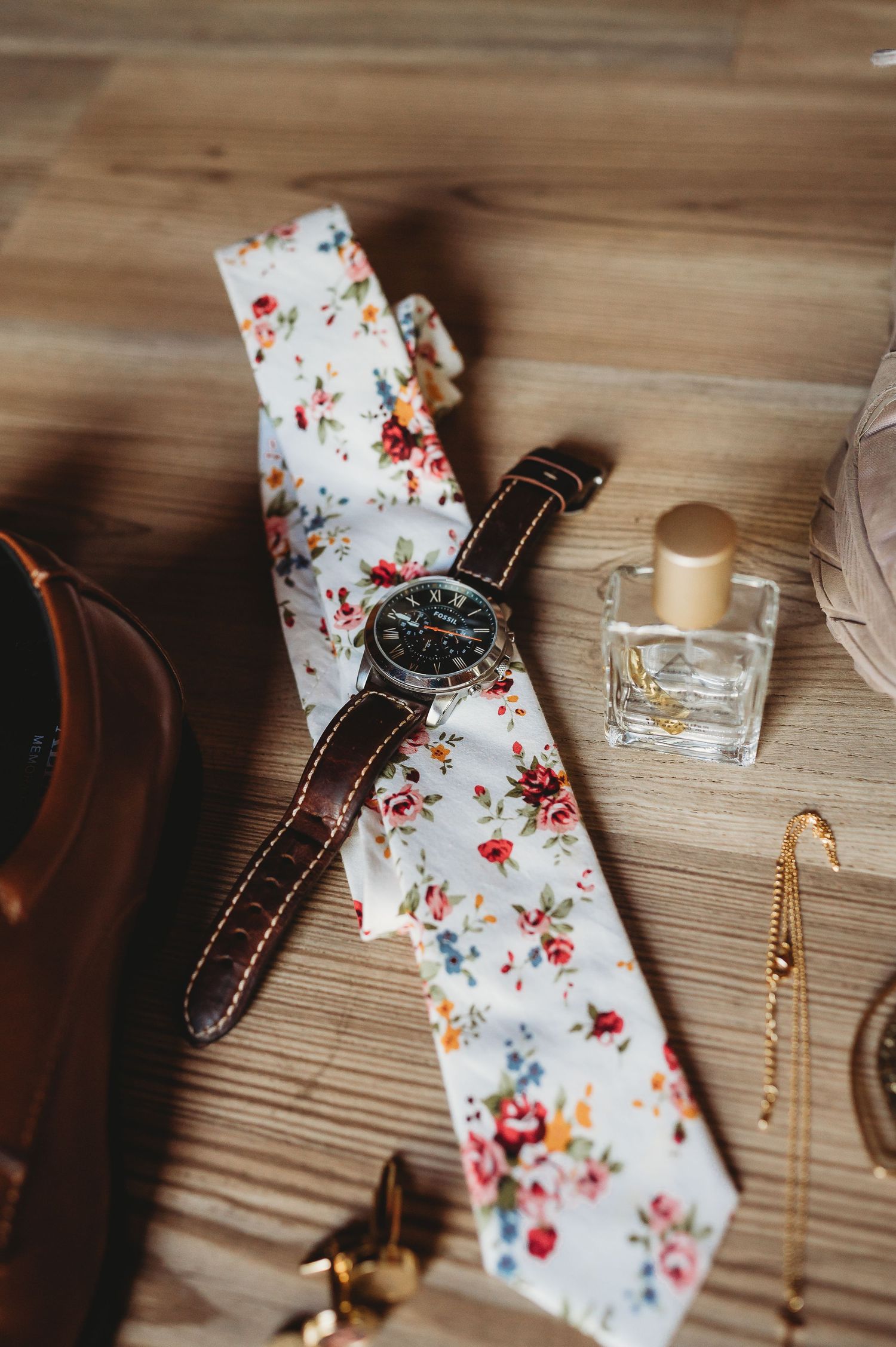 A delicate floral patterned tie and brown leather watch lying on a wooden surface.