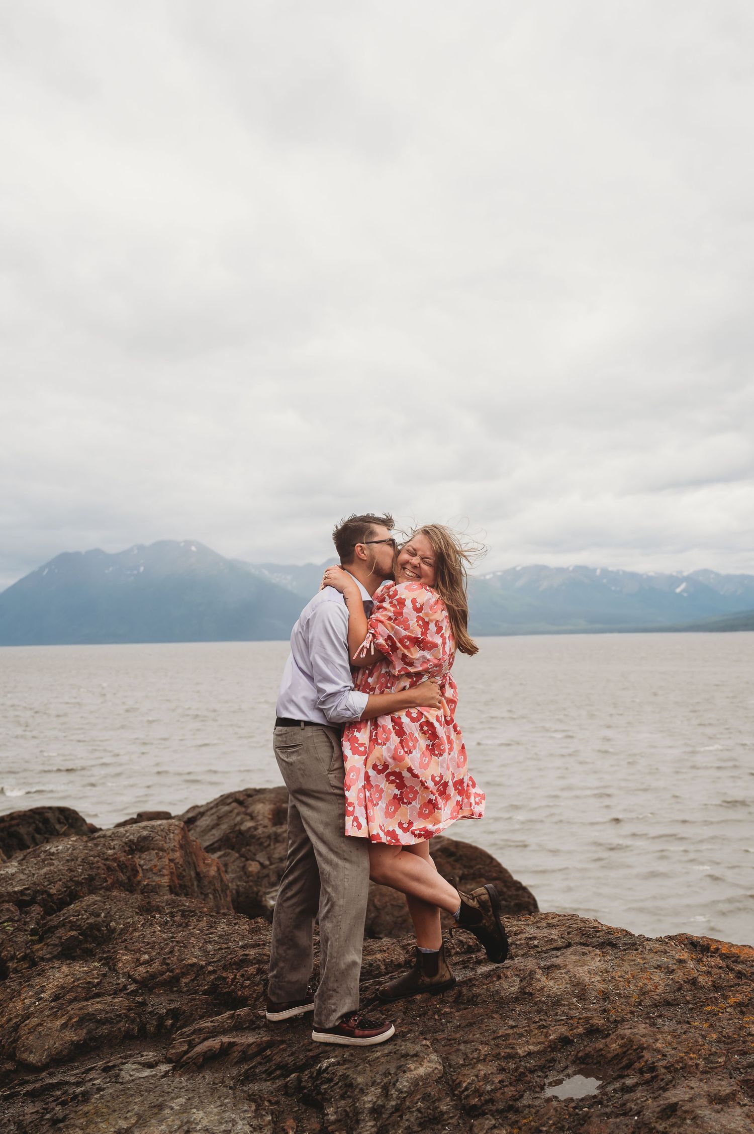 A couple embraces on rocky coastal outcrop with mountains in background under overcast sky.