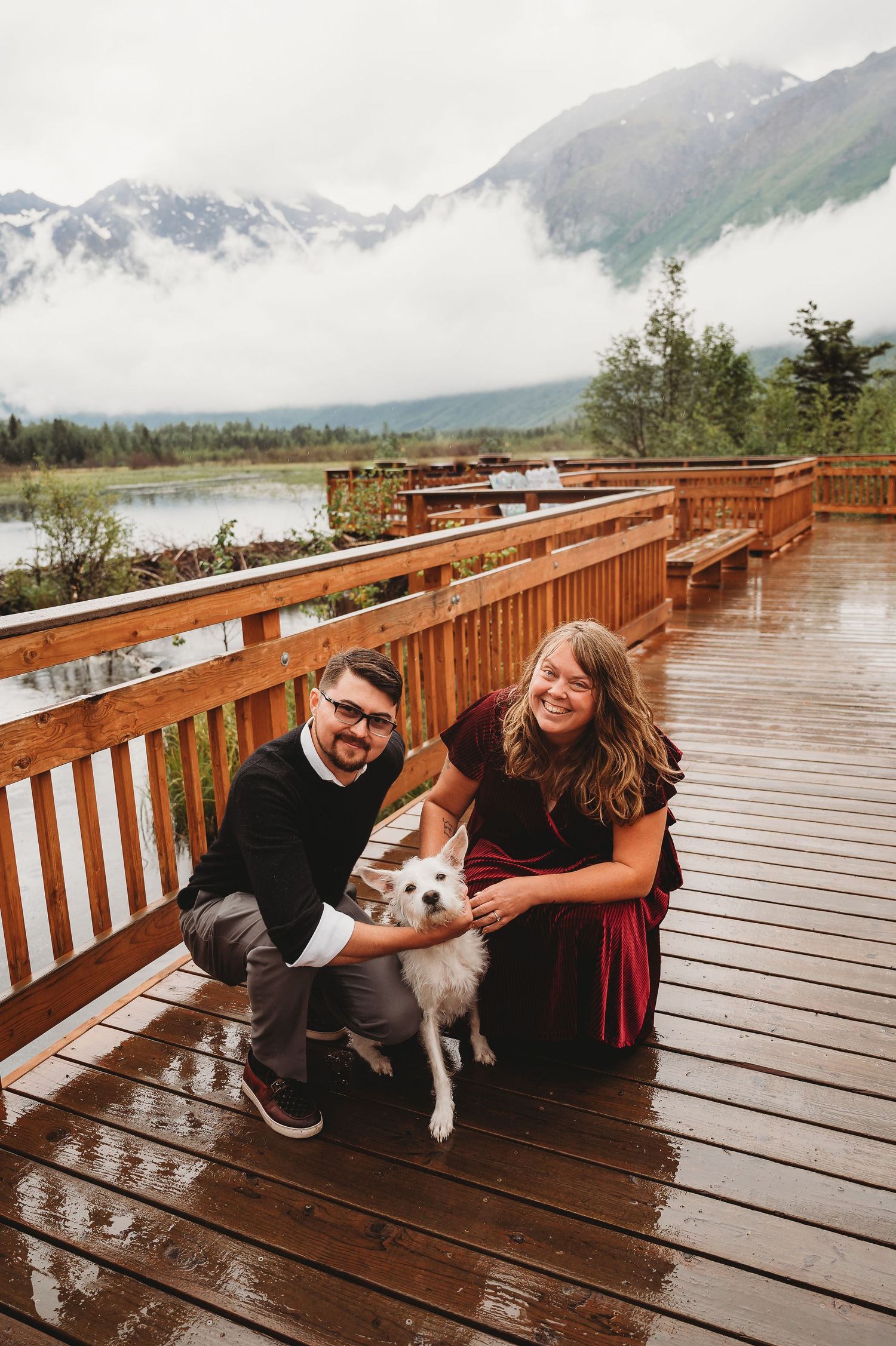 A couple sits with their husky dog on a wet wooden boardwalk overlooking mountain scenery.