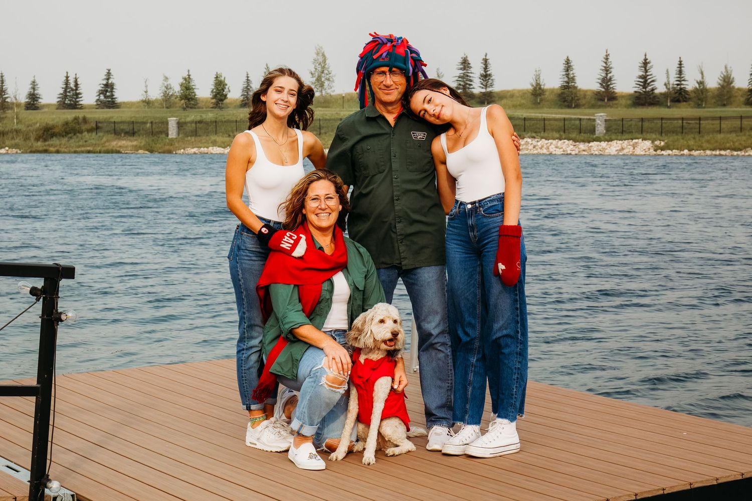A family and their small white dog pose together on a wooden dock by a lake with evergreen trees in the background.