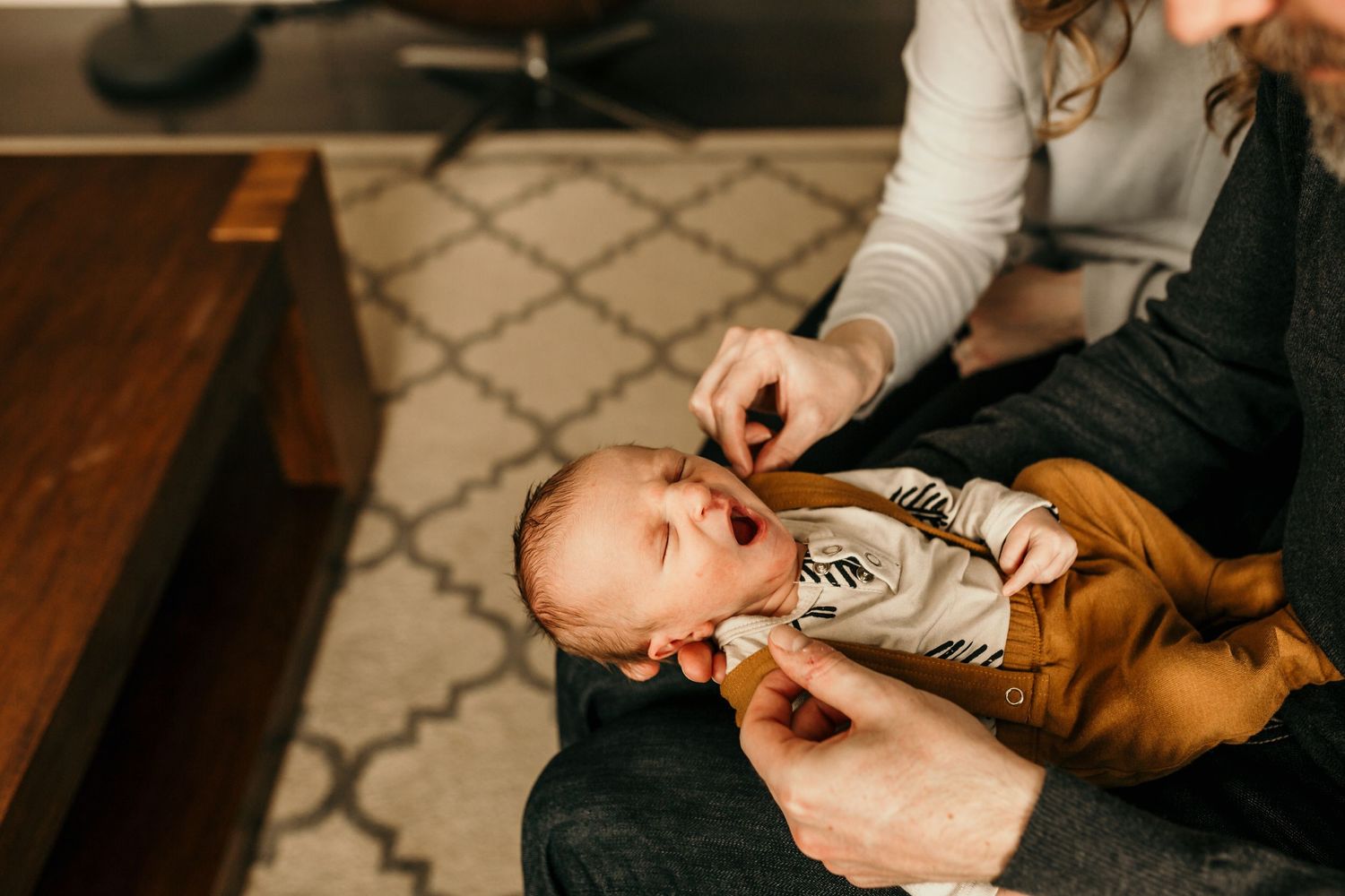 A baby in a brown onesie laughs on a parents lap on a couch over a geometric patterned rug.