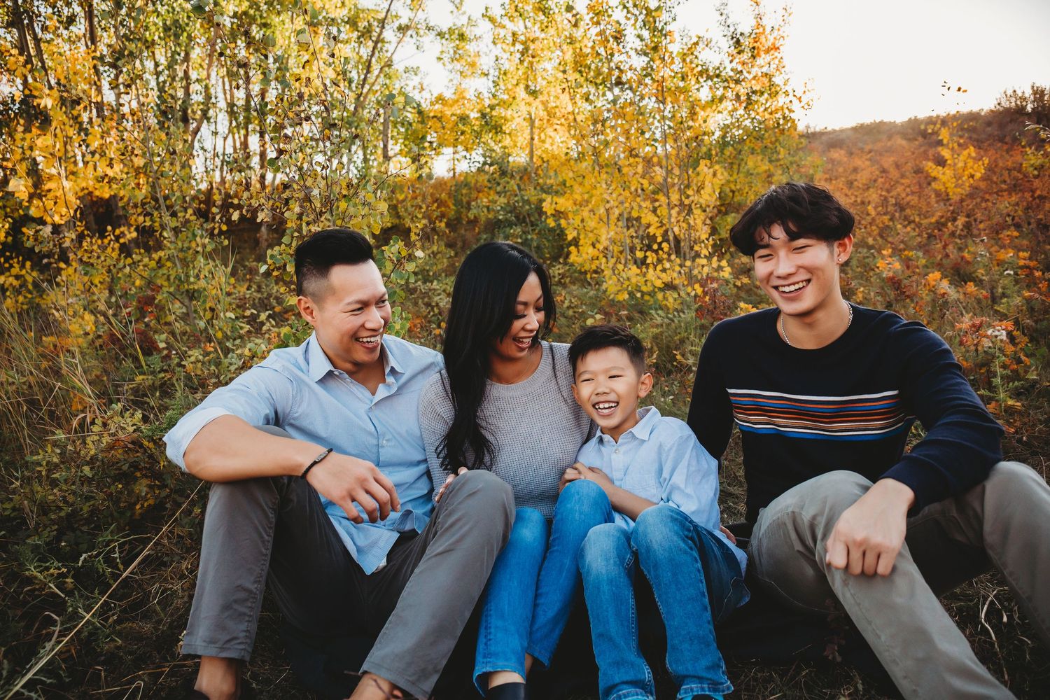 A group sits together outdoors laughing on an autumn day with golden trees and foliage in the background.