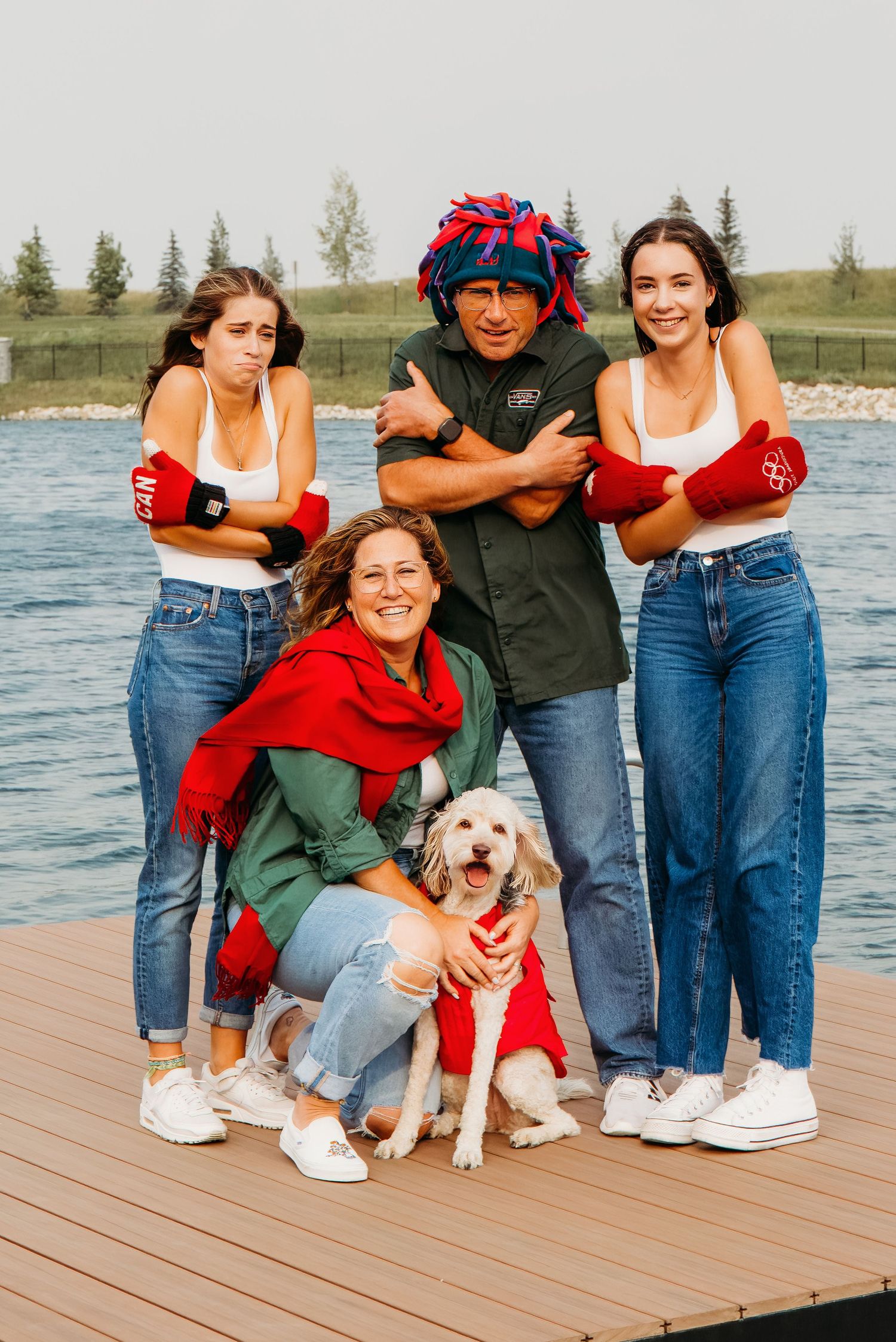 Group stands together on wooden dock by lake wearing casual denim and red outfits with happy golden retriever dog.