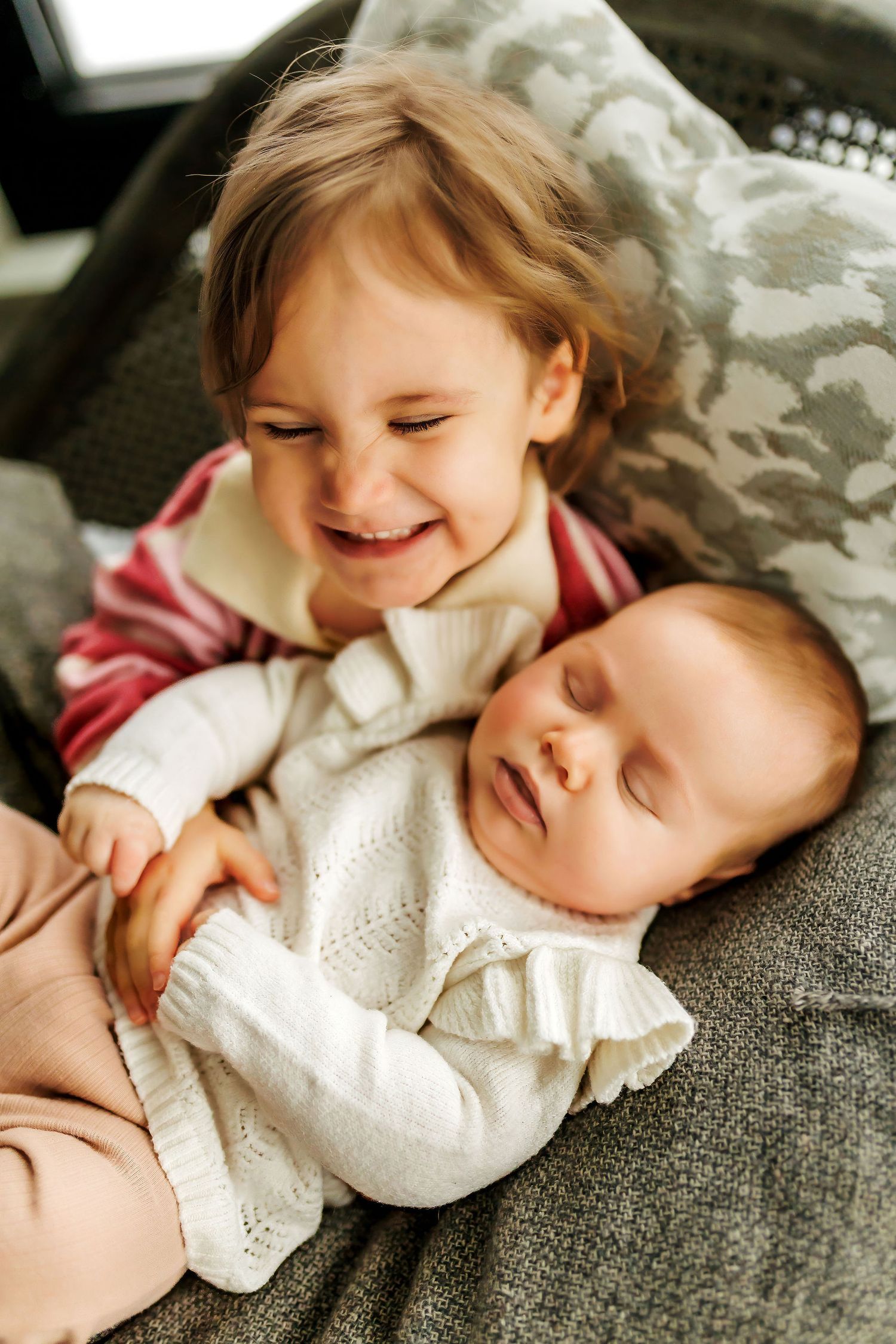Two young children lying together on a grey couch, one smiling while the other sleeps peacefully in a white outfit.