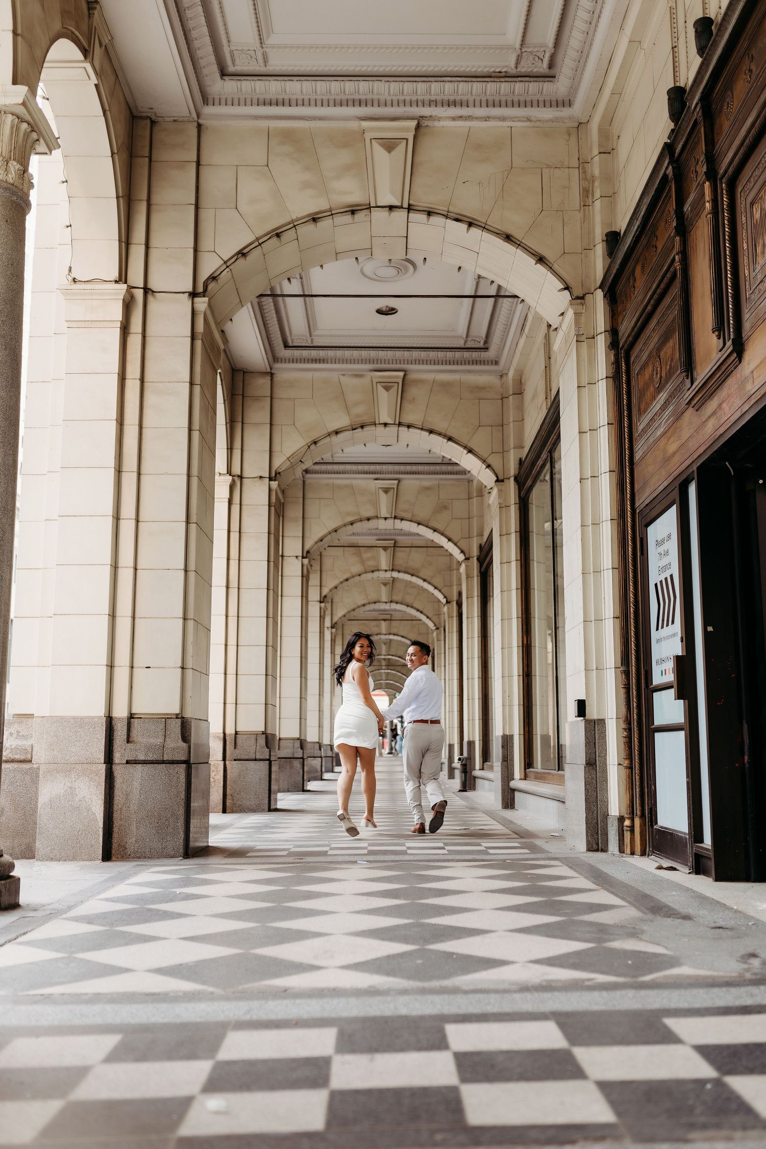 A couple walking through an elegant arched corridor with checkerboard floor in a classical building.