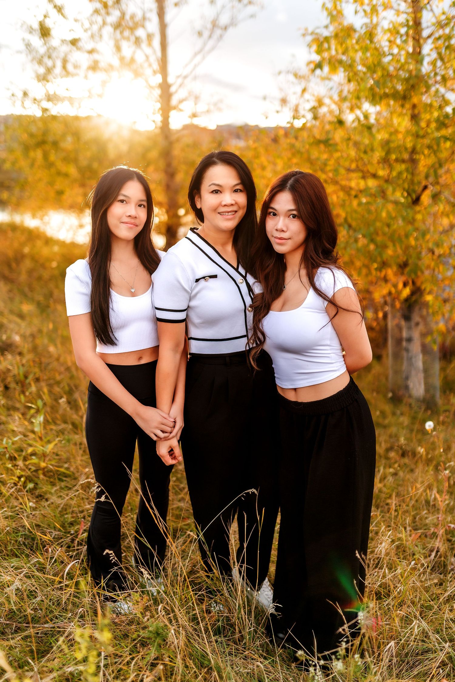 Three women pose together at sunset wearing black pants and white tops in a golden autumn field with trees.