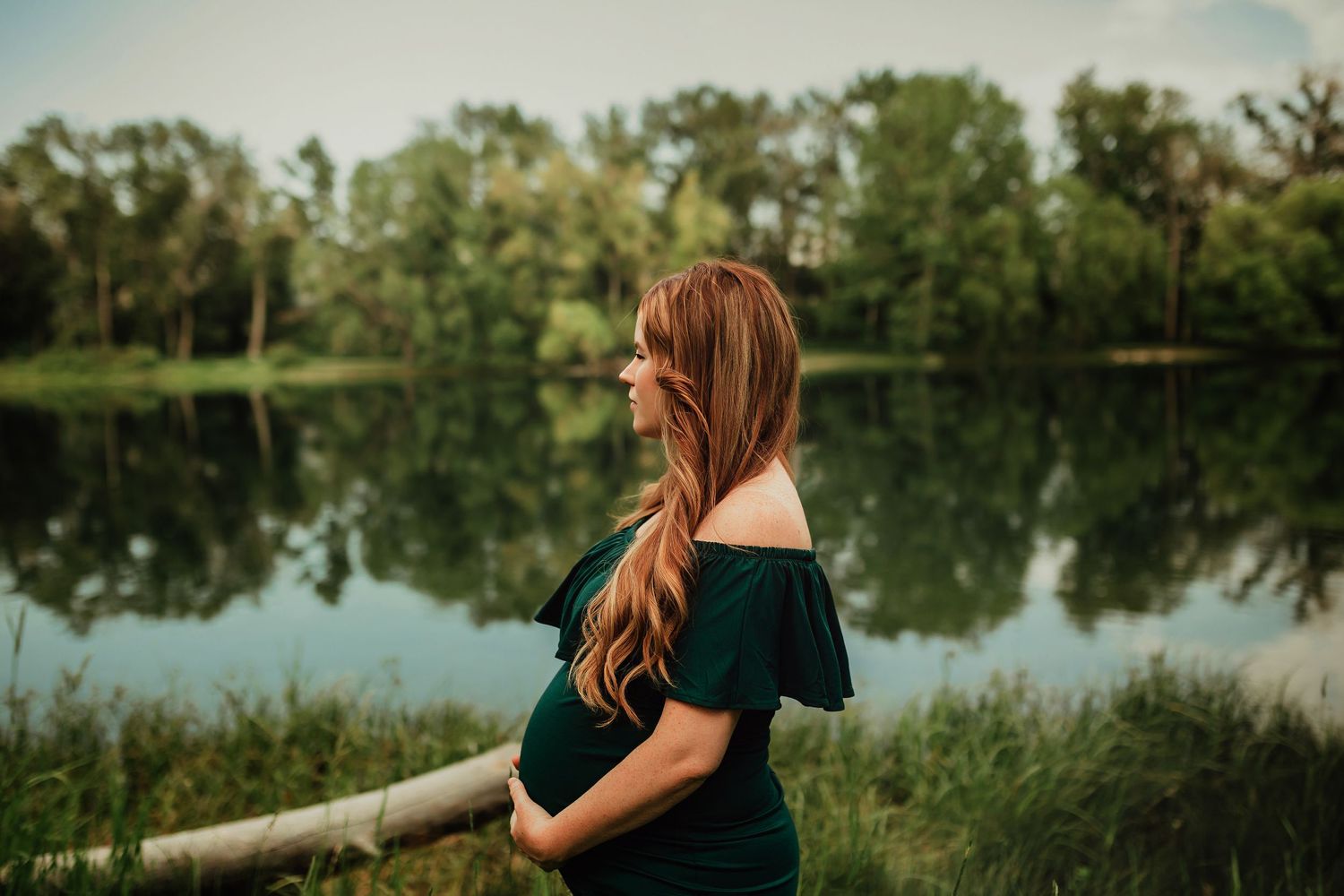 Pregnant woman in black dress stands by calm lake with green trees reflecting in still water at sunset.