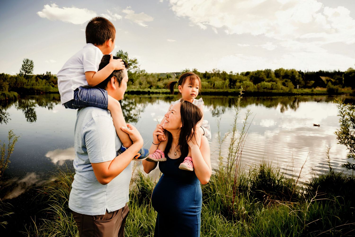 A family enjoys a summer evening by a tranquil lake, with children riding on shoulders against the golden light.