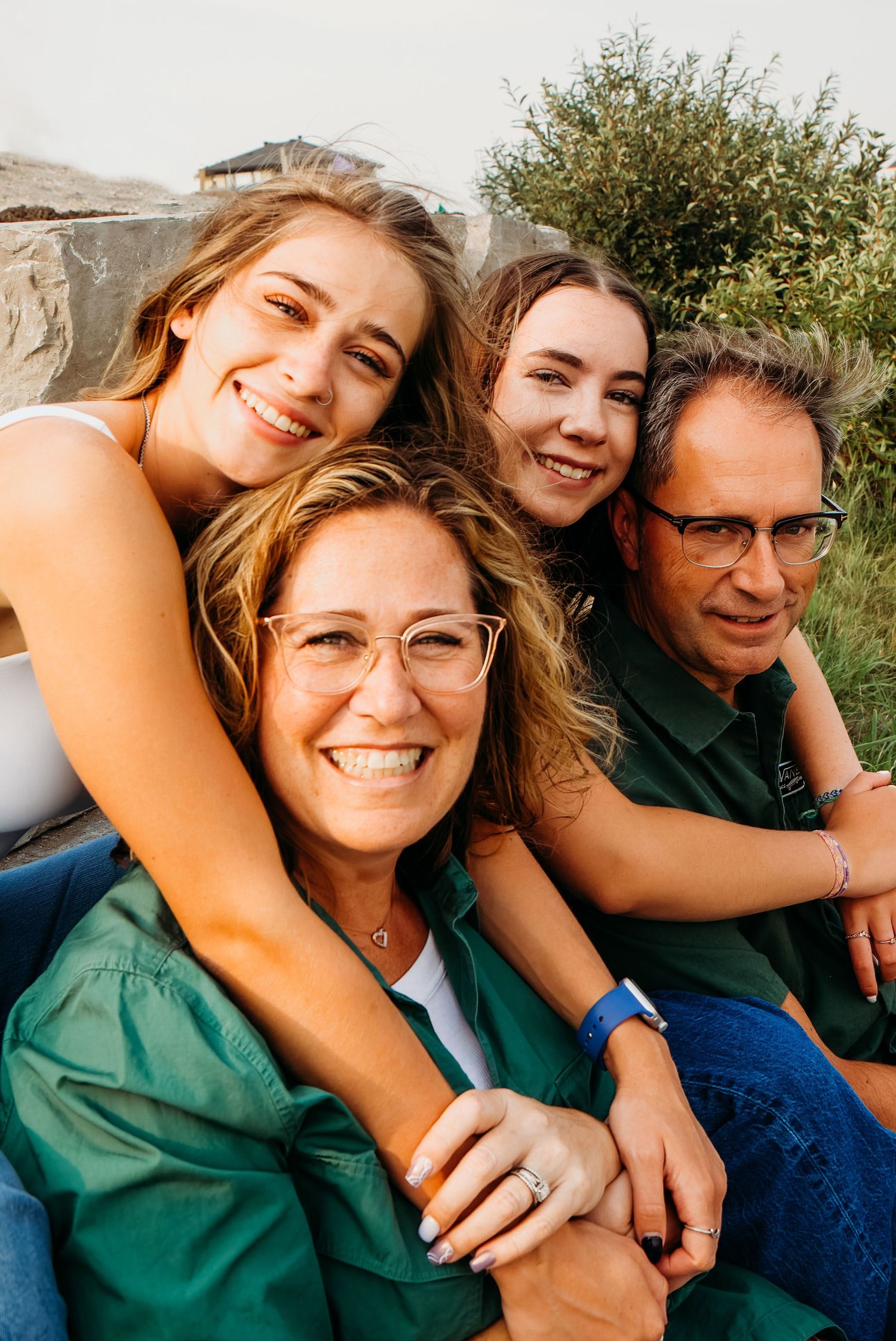 A happy family poses together outdoors in green clothing, smiling warmly at the camera in natural sunlight.