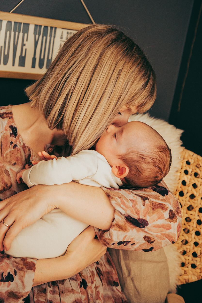 A tender moment between parent and infant in white onesie against warm-toned lighting and wicker decor.