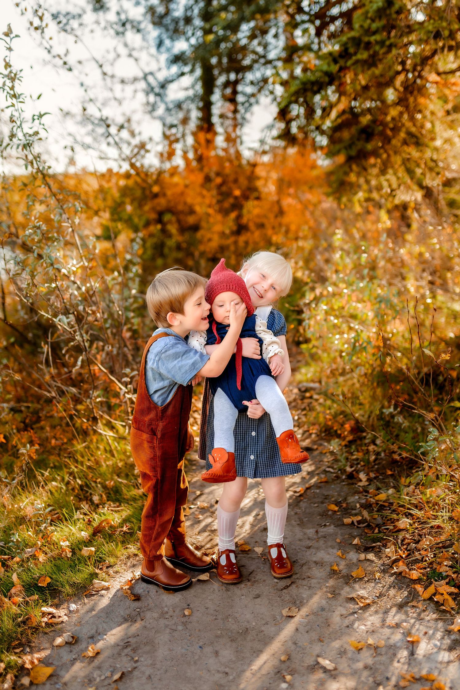 Three children share a playful moment together on an autumn path surrounded by golden foliage.