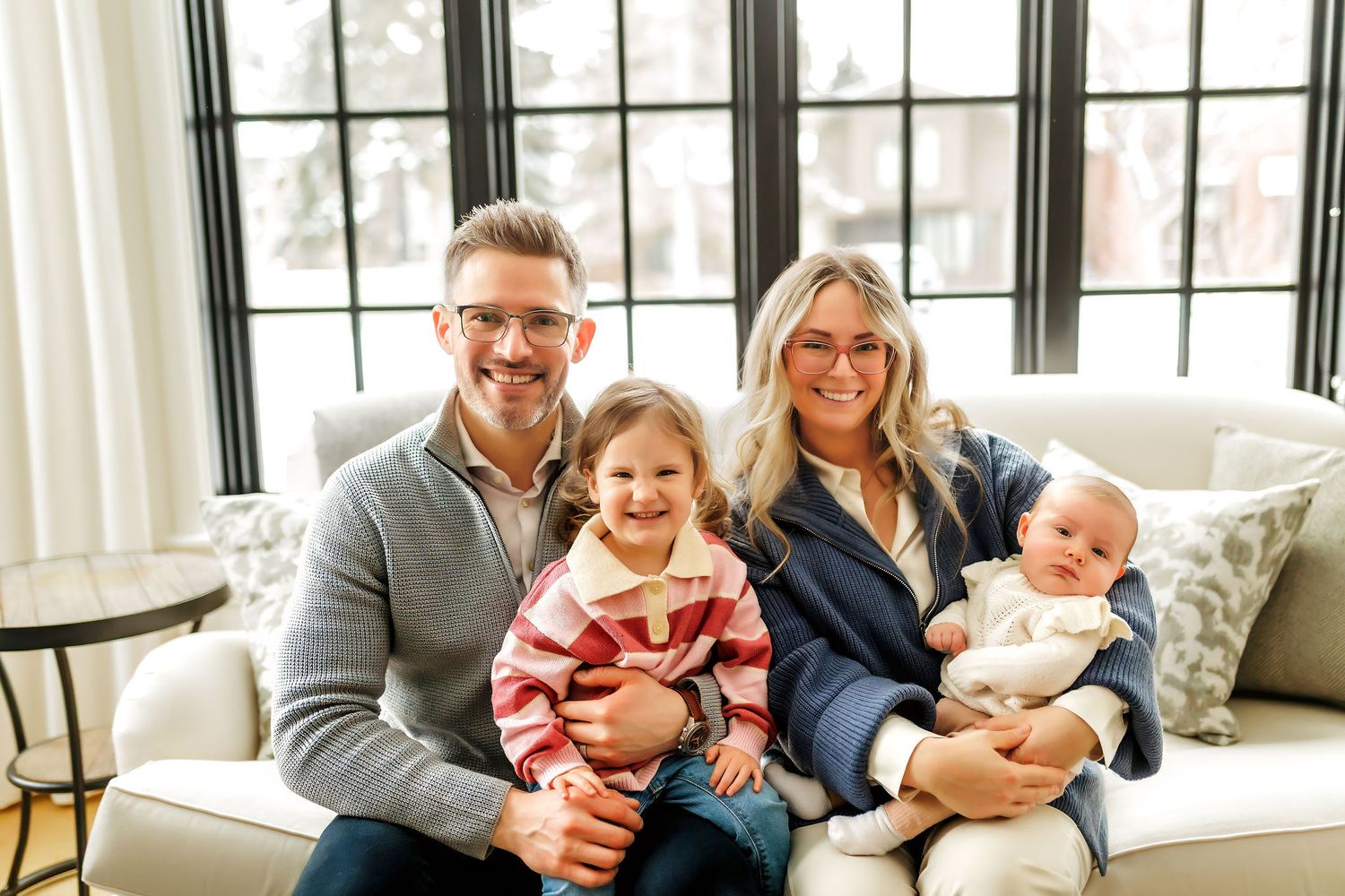 A happy family relaxes together on a couch by a bright window in casual clothing.