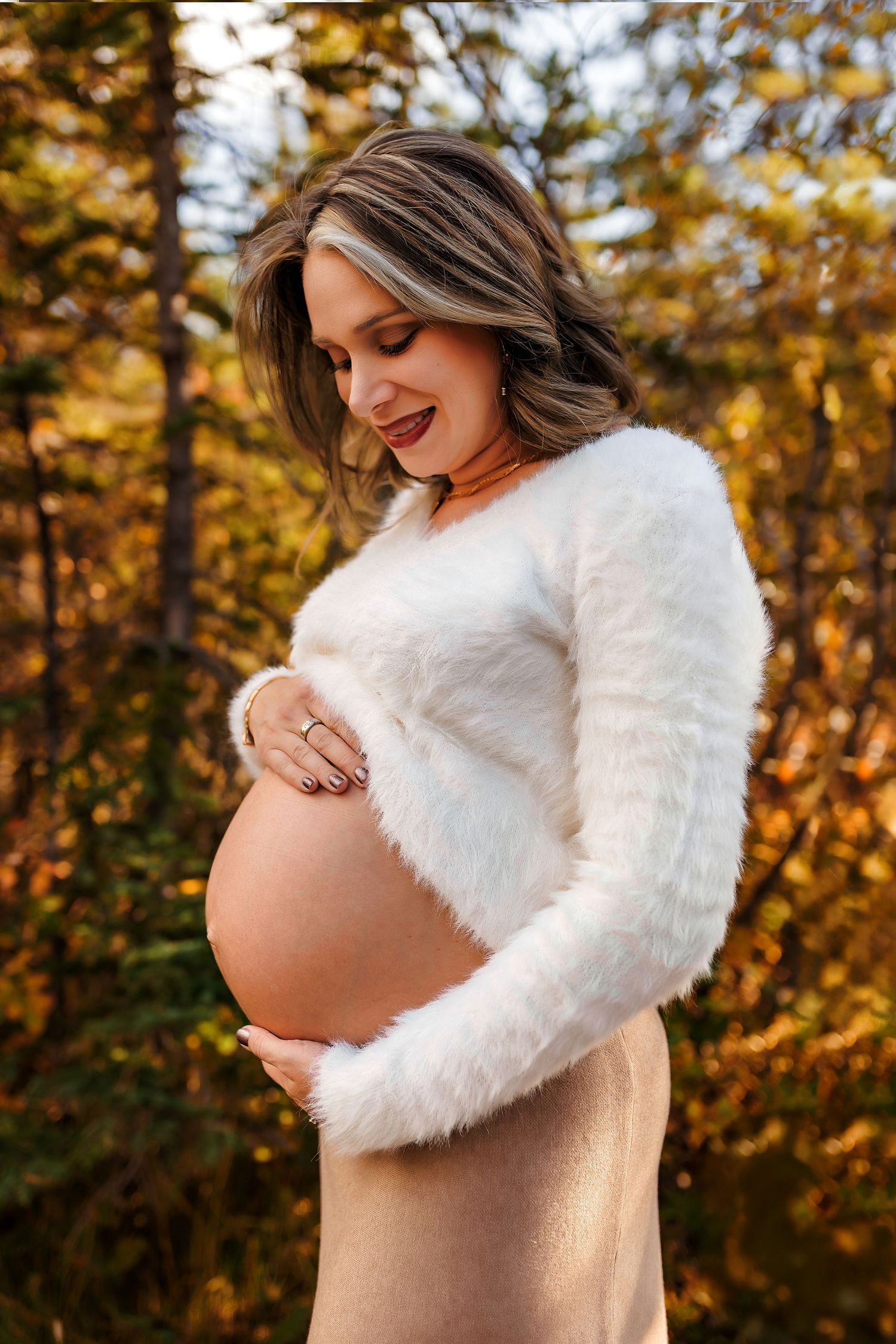A pregnant figure in a white fuzzy sweater poses against golden autumn foliage while gently cradling their belly.