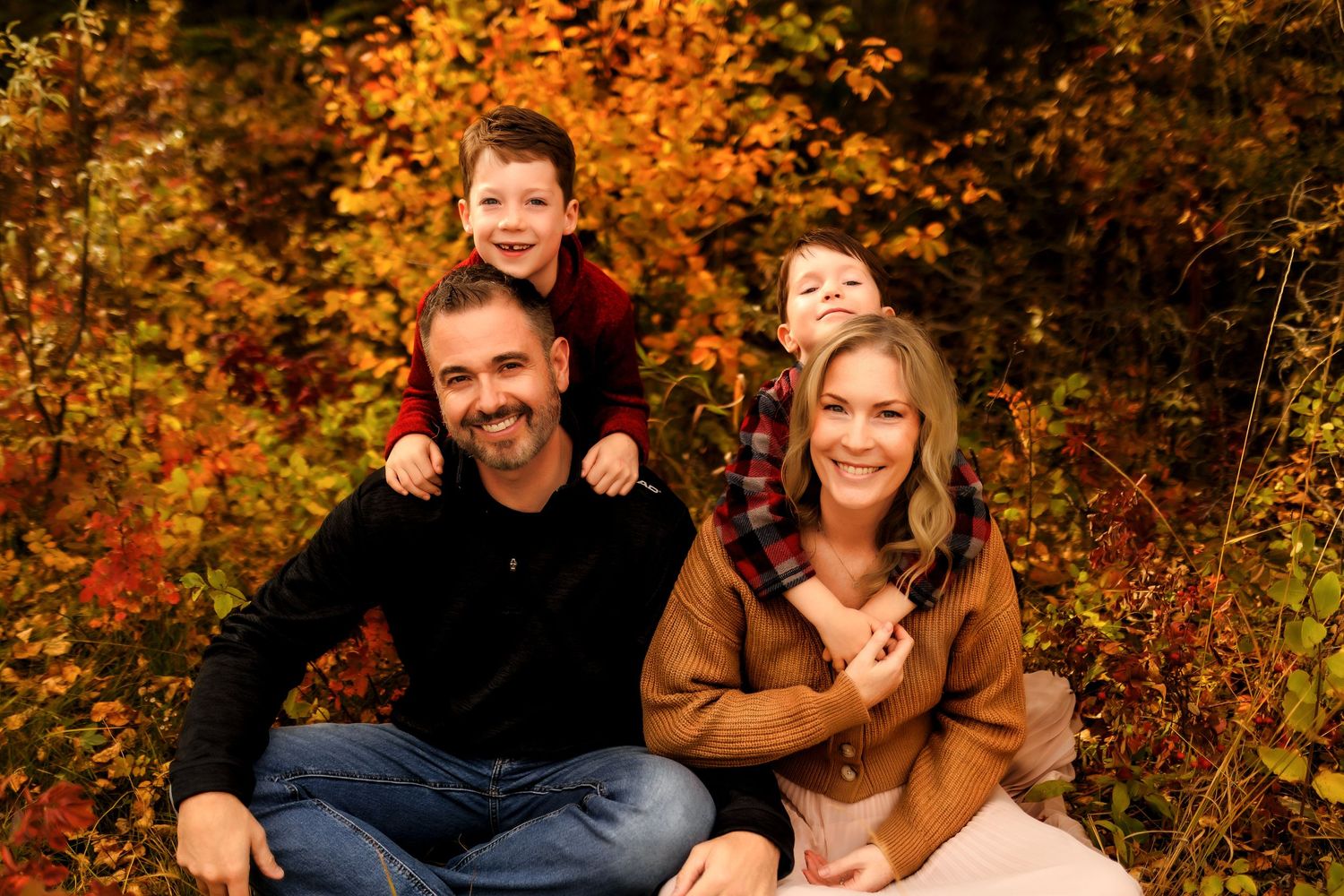 A family wearing fall colors sits together outdoors among vibrant autumn foliage.