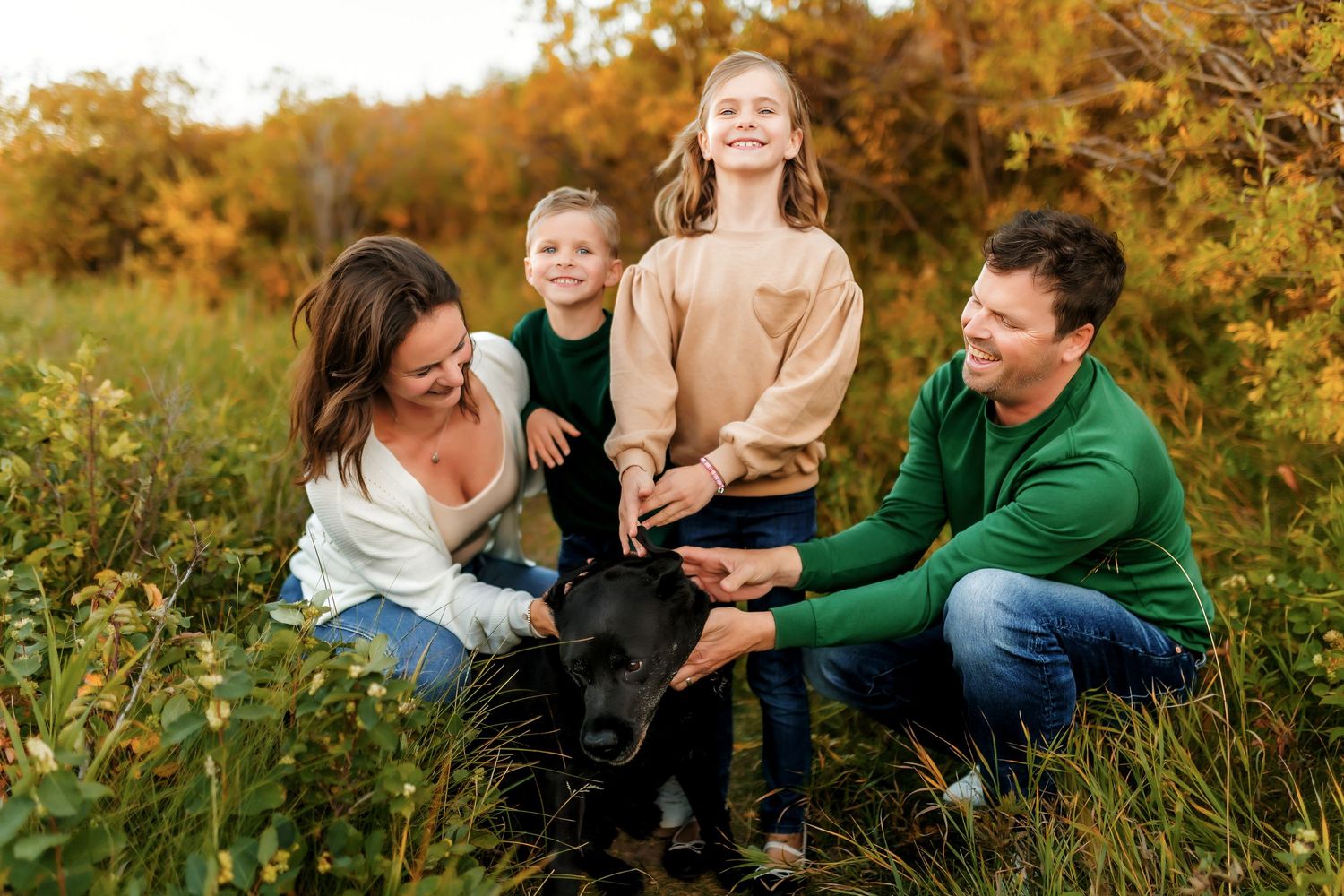 A family plays with their black Labrador dog in a golden autumn field during sunset.