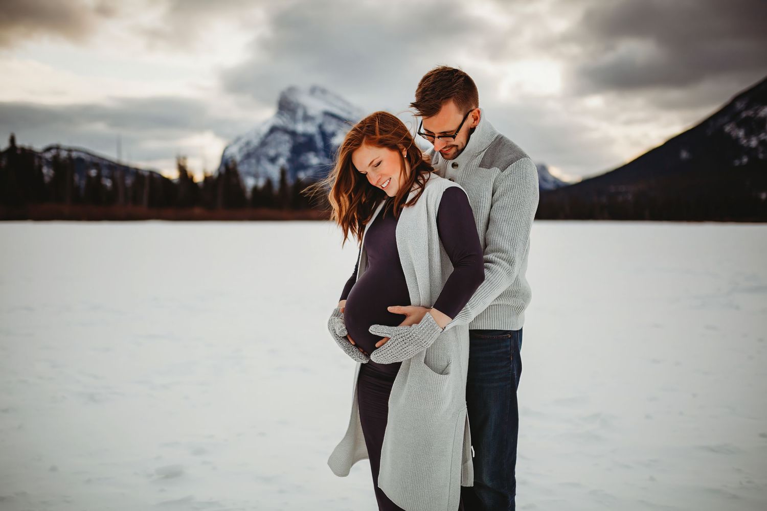 A couple embraces during a winter maternity photoshoot with snowy mountains in the background at Banff National Park.
