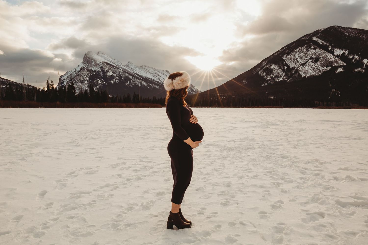 Silhouette figure stands on snowy landscape with dramatic mountain backdrop and sunburst through cloudy sky in Banff.