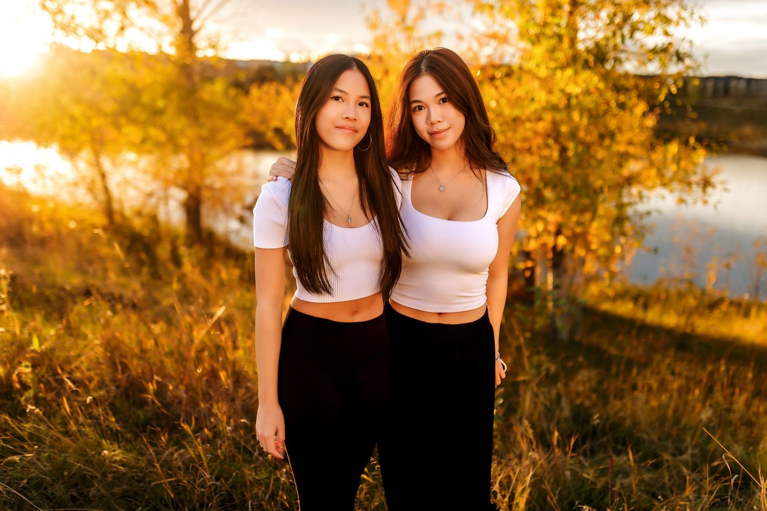 Two women in matching white crop tops and black pants pose outdoors during golden hour sunset near water.