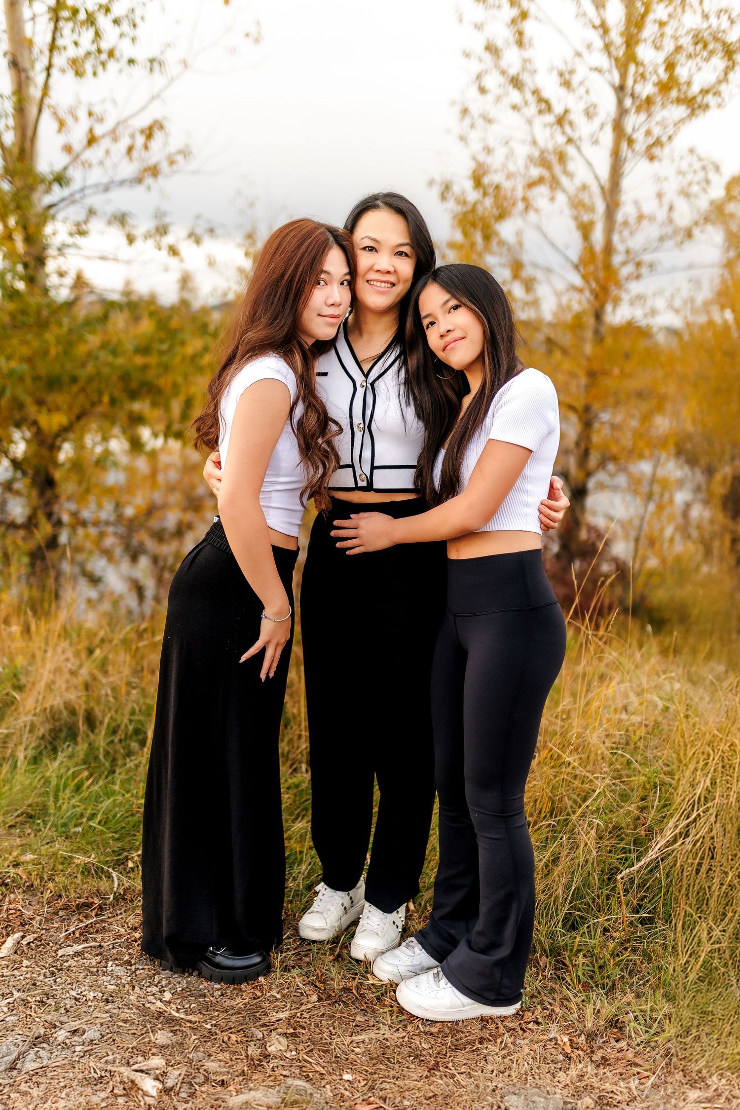 Three friends in casual outfits embracing and smiling together outdoors on an autumn day with golden foliage in background.