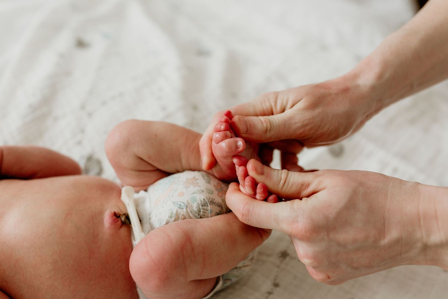 Adult hands gently clean a newborn baby's tiny feet on white fabric.