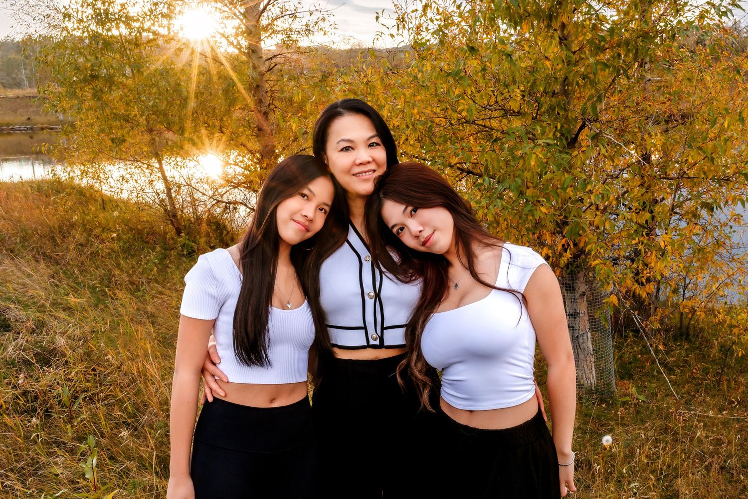 Three friends in crop tops pose together at sunset by a lake with autumn foliage in the background.