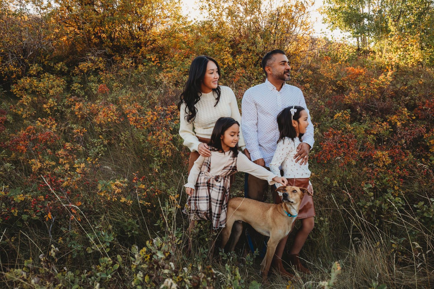 A family wearing coordinated white and beige outfits poses together in an autumn field with golden foliage.