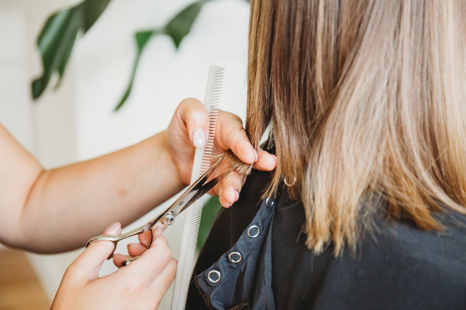 Professional hairstylist using scissors to trim blonde hair ends while wearing a black salon cape.