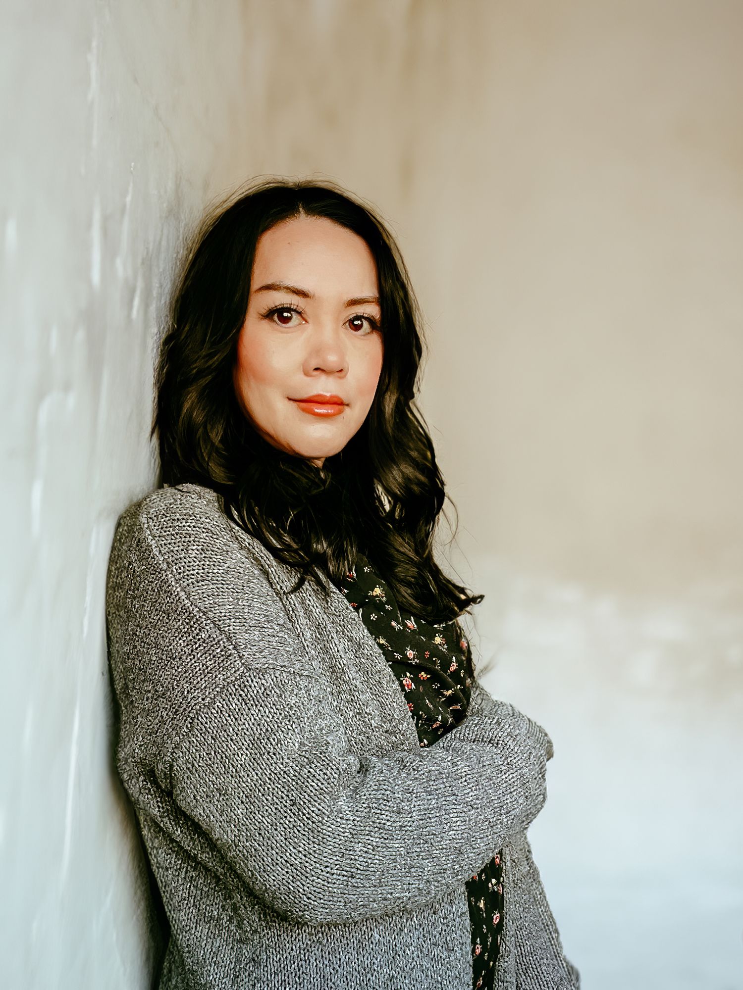 Woman in gray cardigan sweater and floral top poses against white wall for a stylish profile portrait.