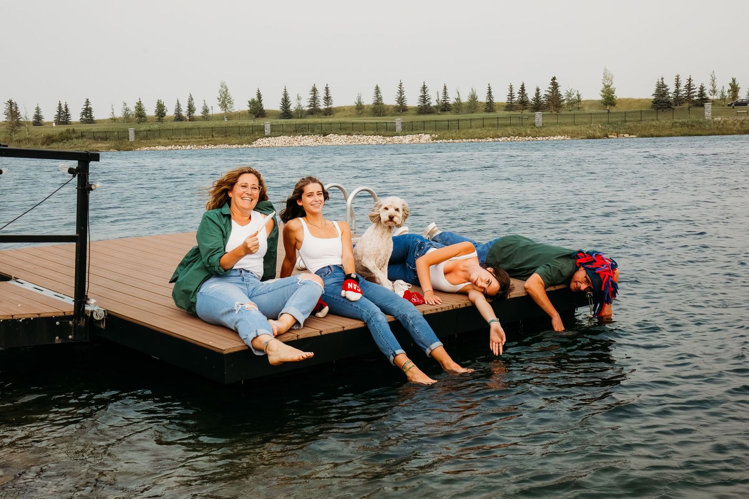 Four people relax and laugh together while lounging on a wooden dock by a lake with evergreen trees in the background.