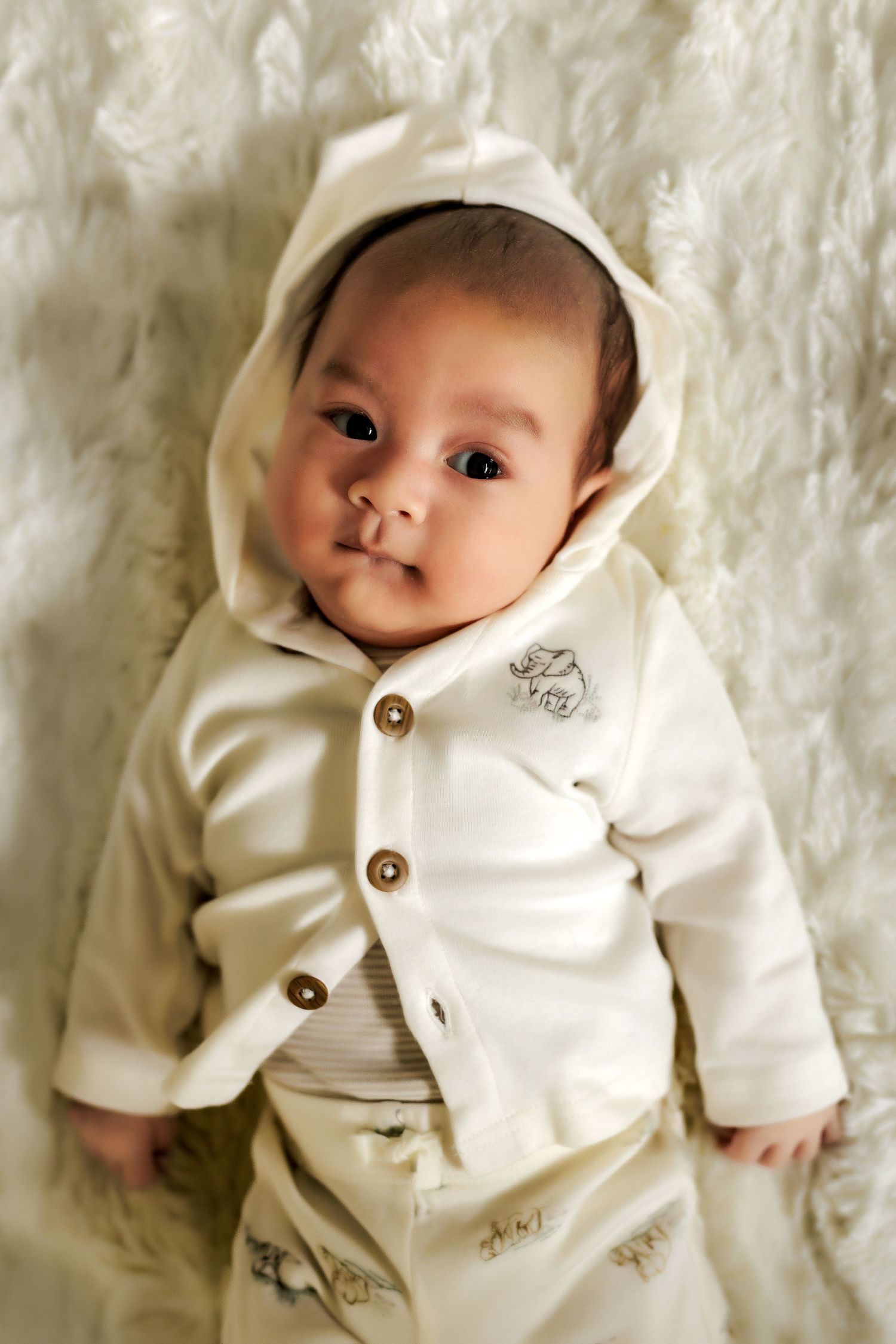 Adorable baby wearing a cream-colored hooded sweater with buttons lies on soft white textured fabric.