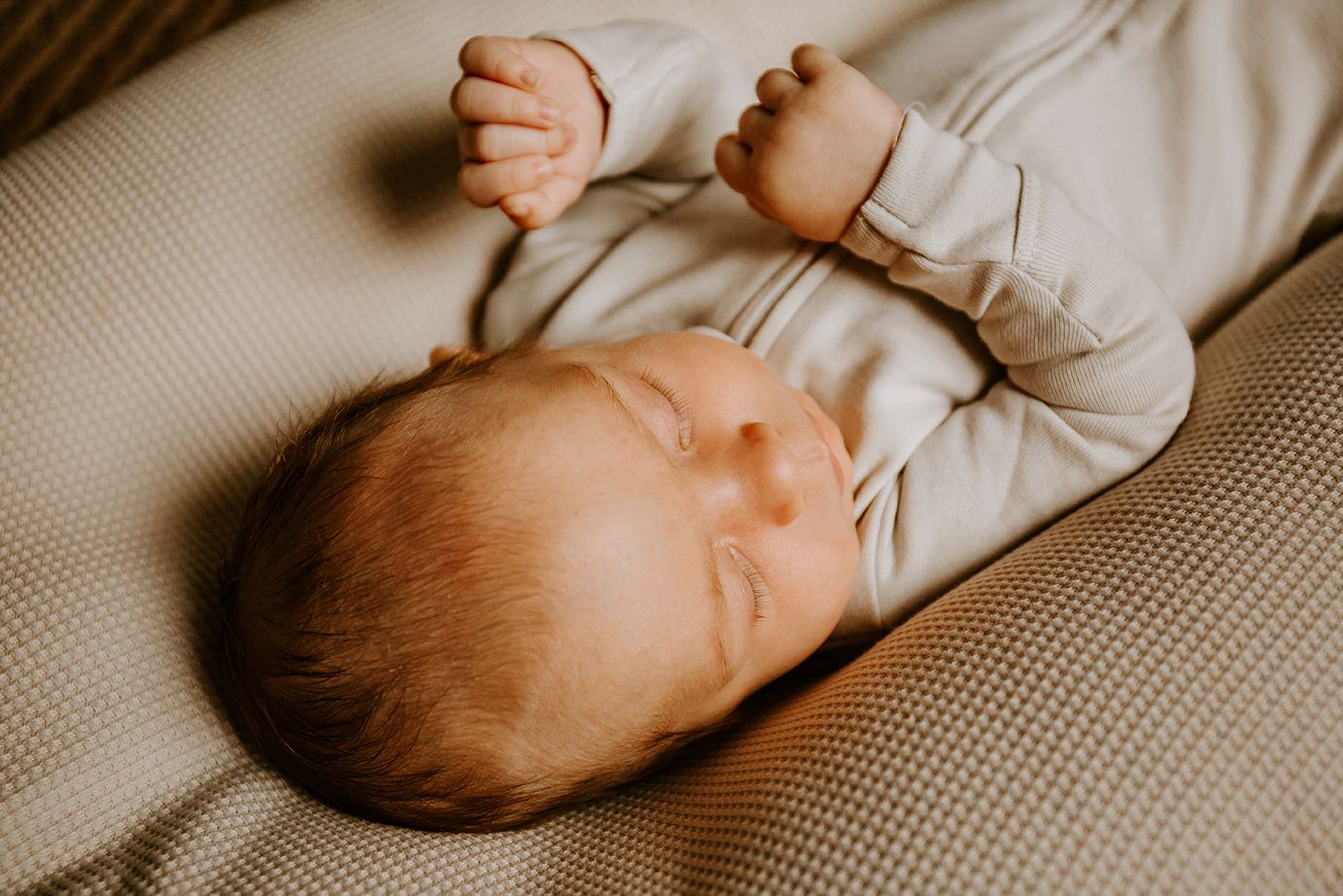 A baby wearing light gray clothing stretches while resting on textured fabric bedding.