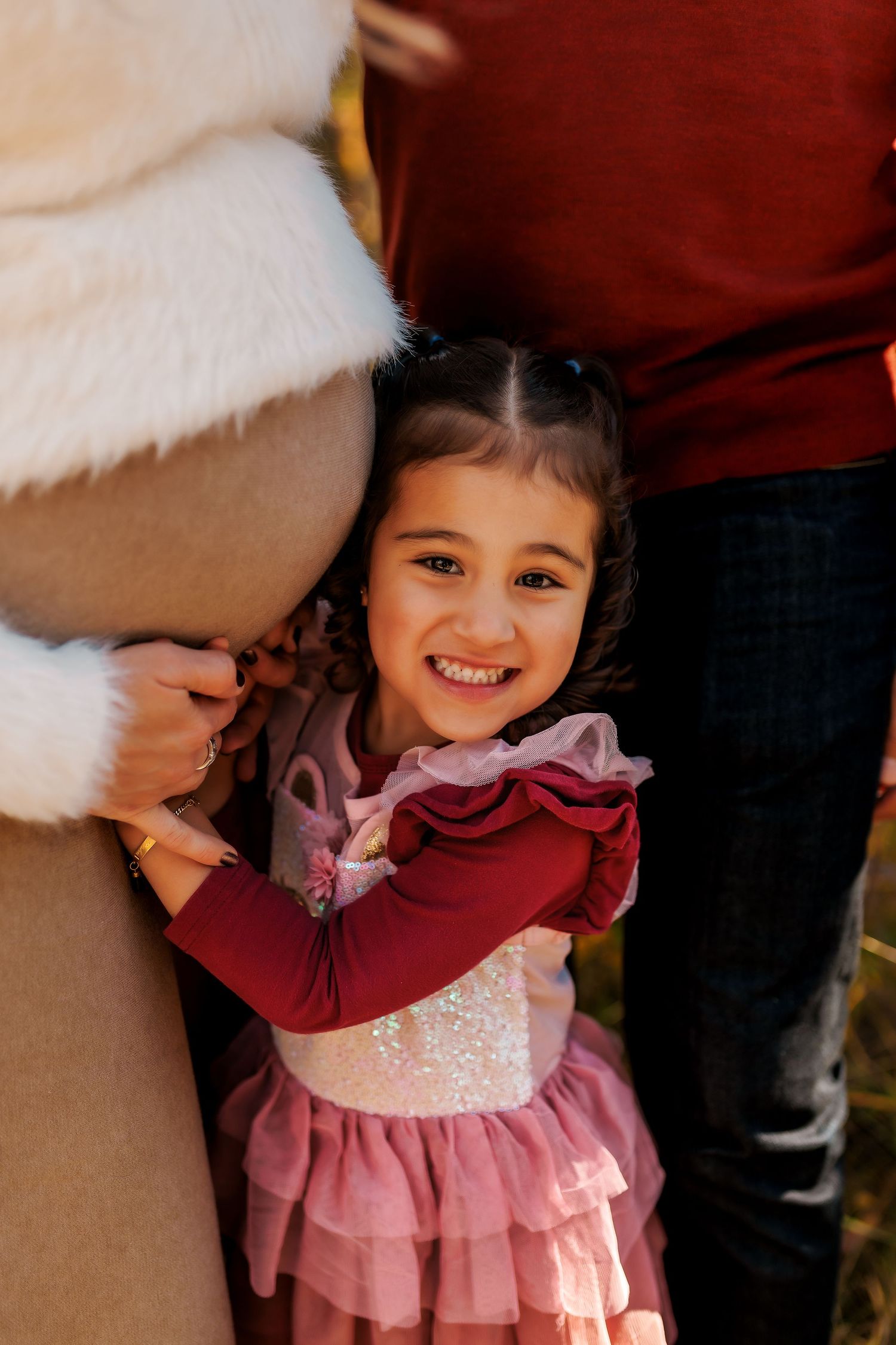 A child in a pink ruffled dress stands next to someone in a beige coat with white fur trim during what appears to be winter.
