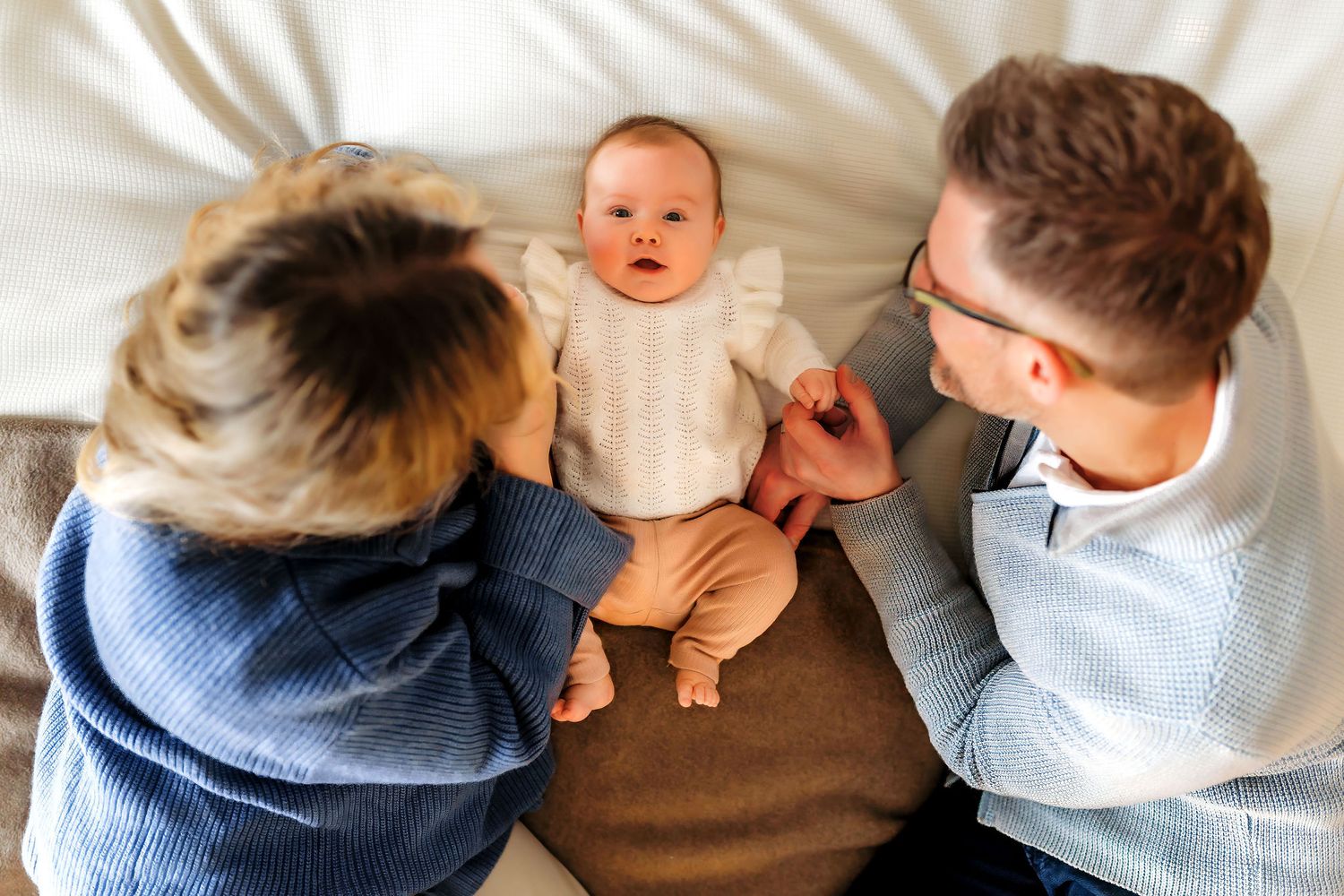 A small baby lies on a brown blanket while people on either side play and interact.