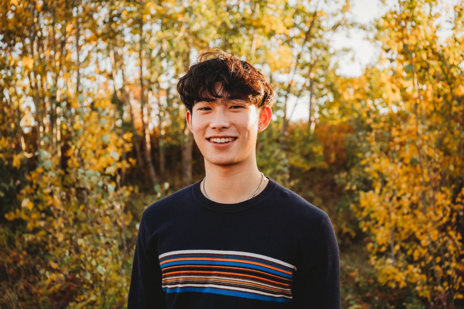 Portrait of someone in a striped sweater smiling outdoors with autumn foliage and golden trees in the background.