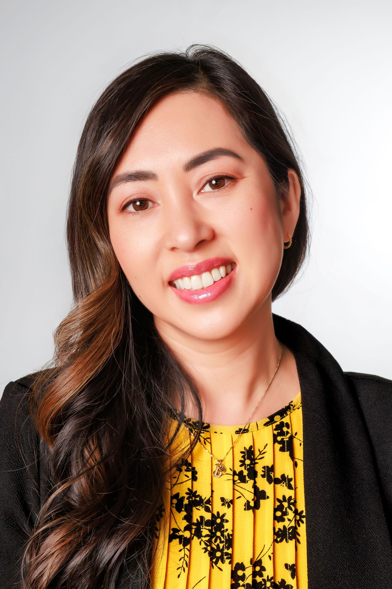 Professional headshot with long dark hair wearing a black blazer and yellow floral top against light background.
