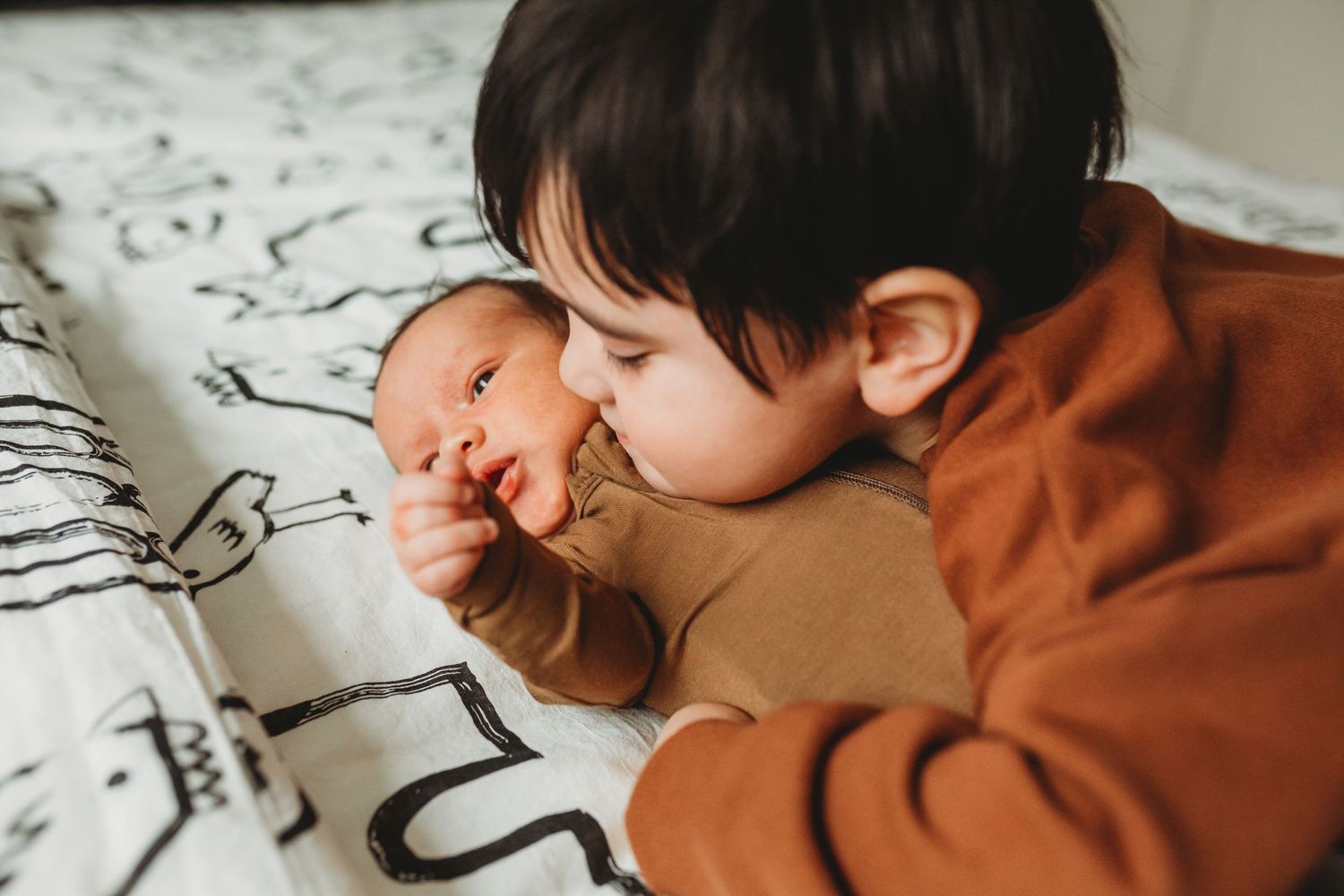 Two siblings lying together on a patterned blanket as the older one in an orange sweater shows affection to the younger one.