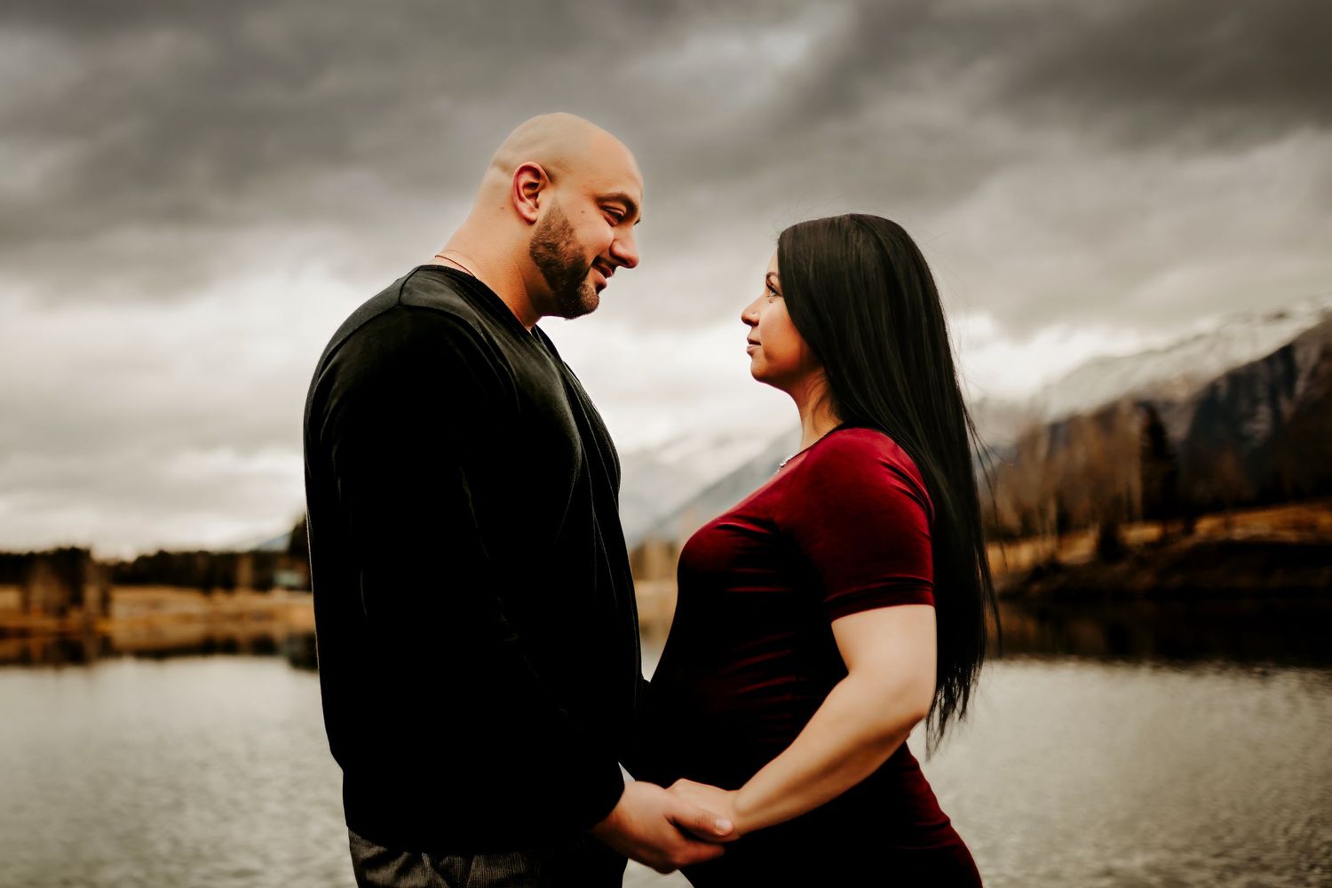 A couple in dramatic lighting shares an intimate moment by a serene lake with mountains and stormy skies in the background.