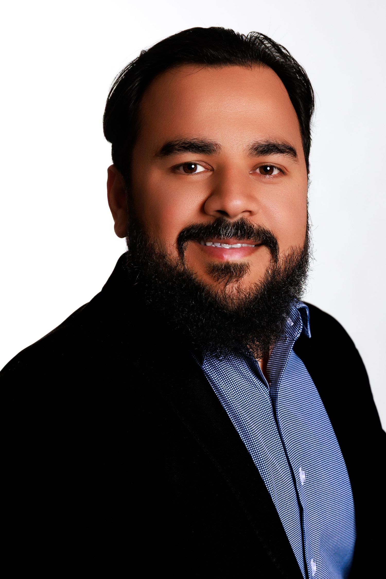 Professional headshot of person wearing blue collared shirt against white background smiling warmly with full beard.