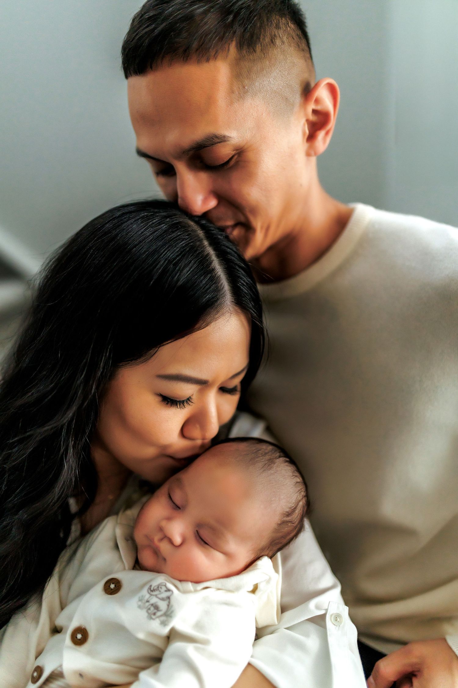 A caring couple shares an intimate moment while holding their sleeping newborn baby in a warm, natural light setting.