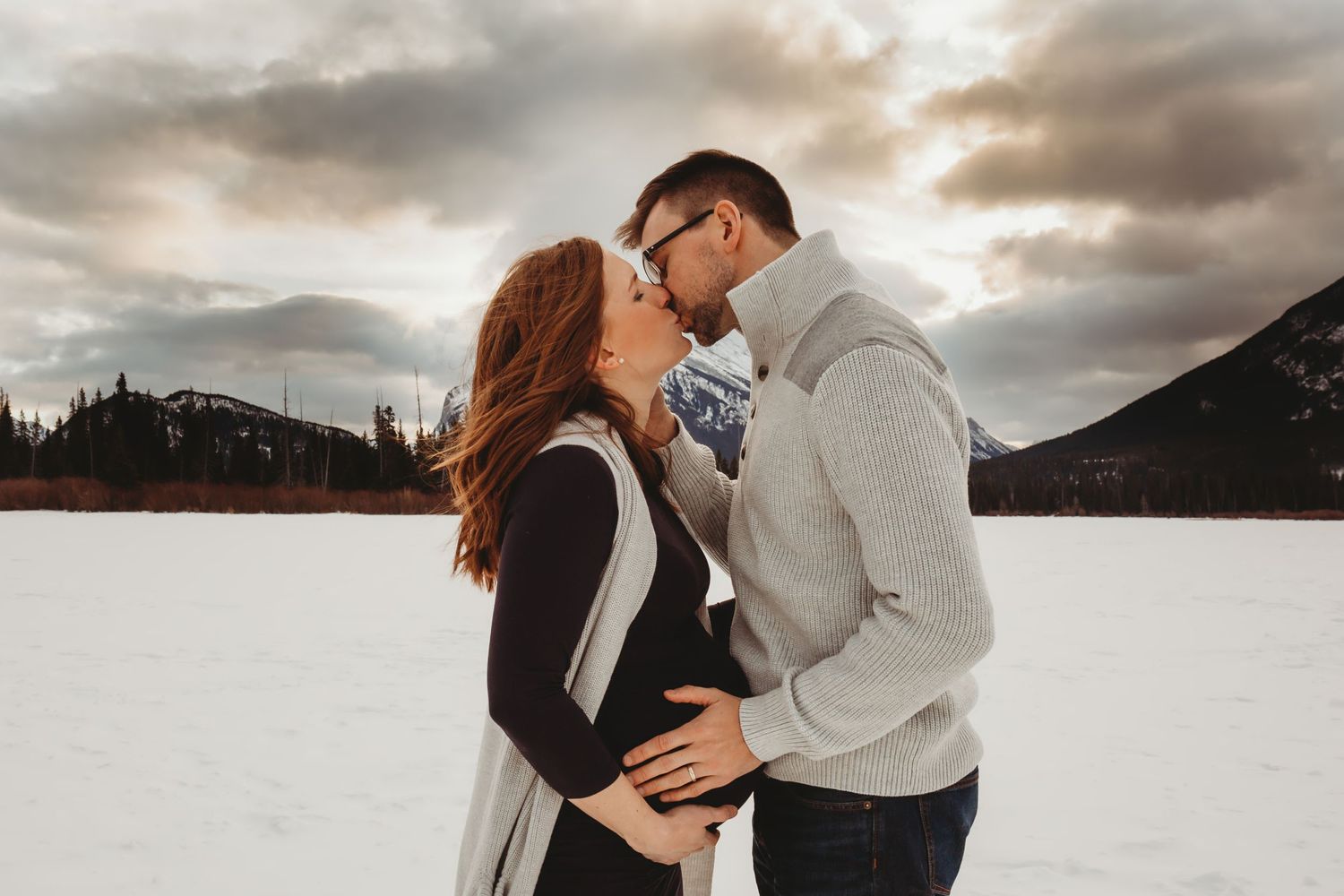 A romantic winter engagement photo with a couple kissing in front of snowy mountains and dramatic cloudy skies.
