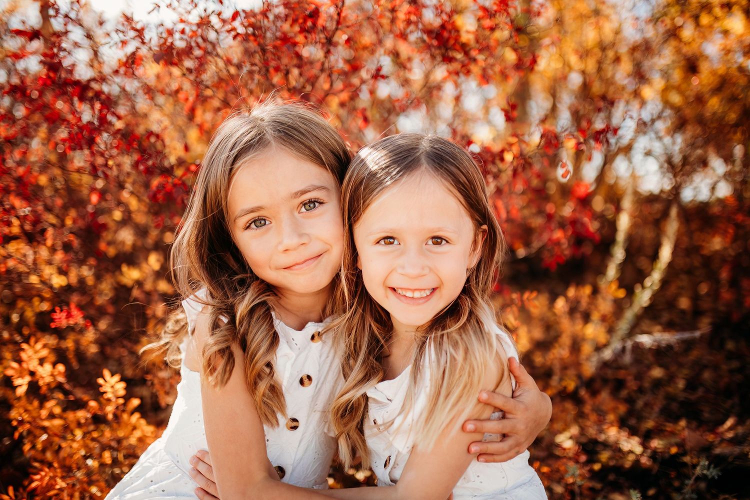 Two children in white outfits smile together against vibrant orange and red autumn foliage in background.