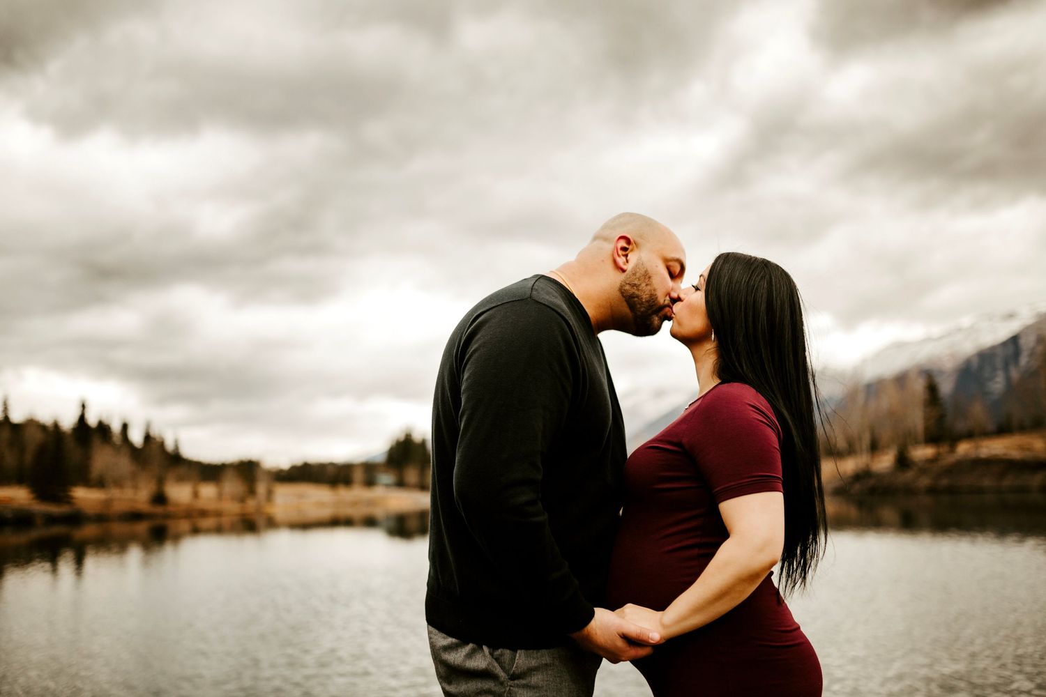Couple sharing a romantic moment by a serene mountain lake under dramatic cloudy skies.
