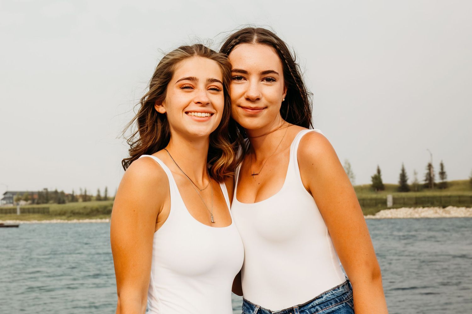 Two women in white tank tops pose together and smile by a lake on a sunny summer day.