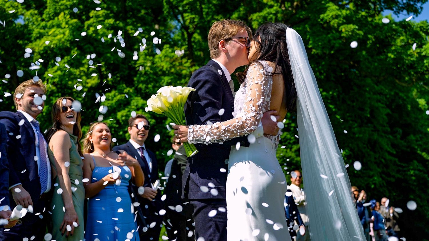 A newlywed couple kisses during their outdoor garden ceremony while guests celebrate with confetti and flowers.