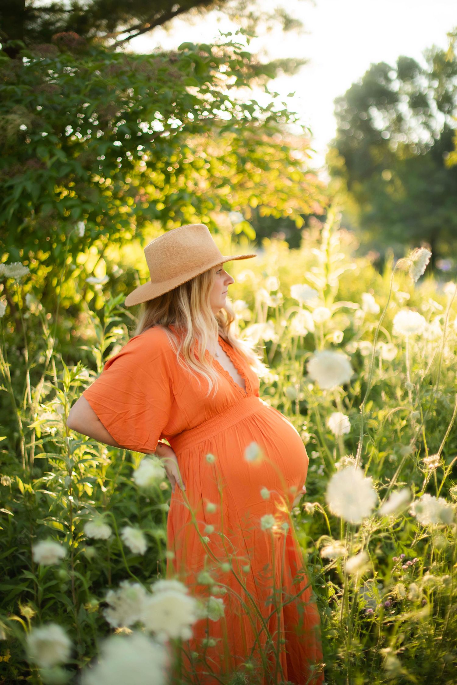 Pregnant woman in orange dress and straw hat standing among wildflowers in golden sunlight.