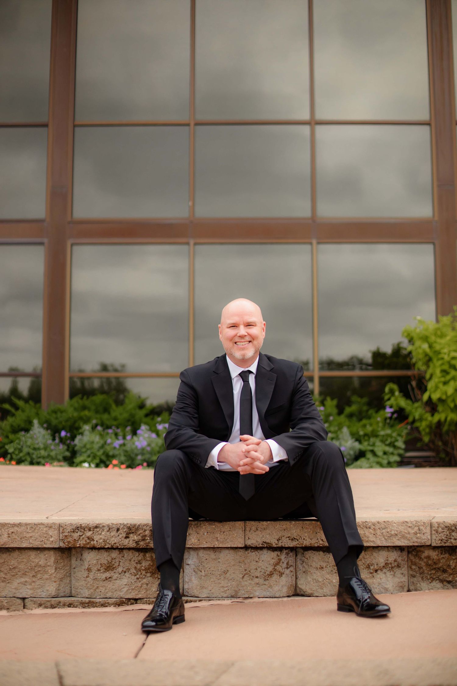 Professional portrait of someone in a dark suit sitting casually on stone steps in front of a modern glass building.
